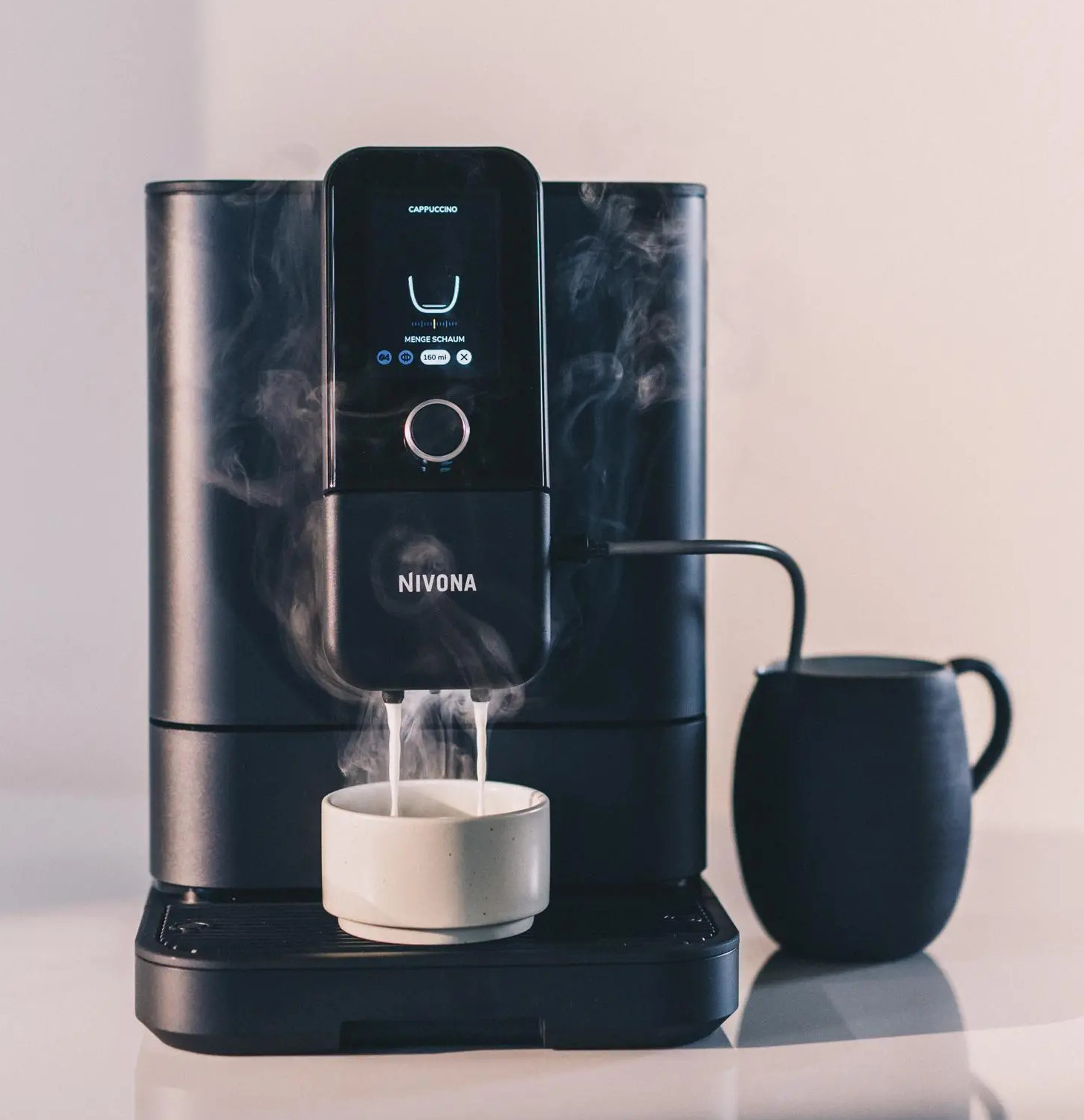 Czarny ekspres do kawy Nivona wydziela parę do filiżanki. Po prawej stronie stoi czarny kubek. Na ekranie napis 'Cappuccino'.