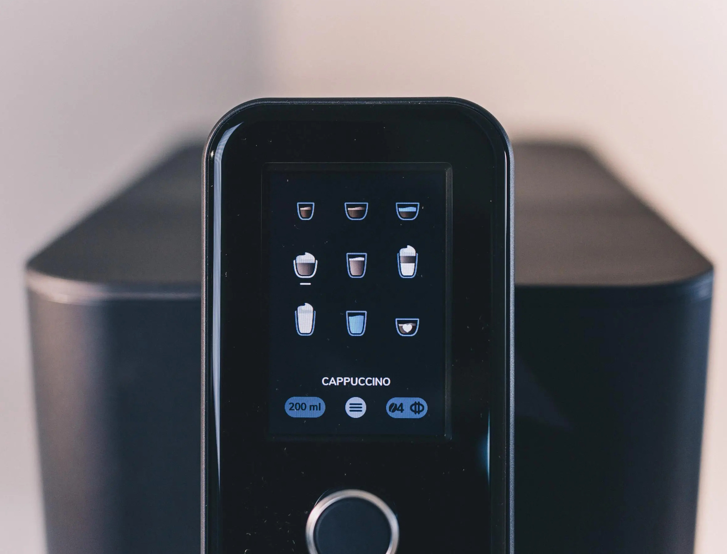 Wyświetlacz ekspresu do kawy pokazuje opcje cappuccino. Czarna maszyna z ekranem i przyciskami. Beżowe tło.