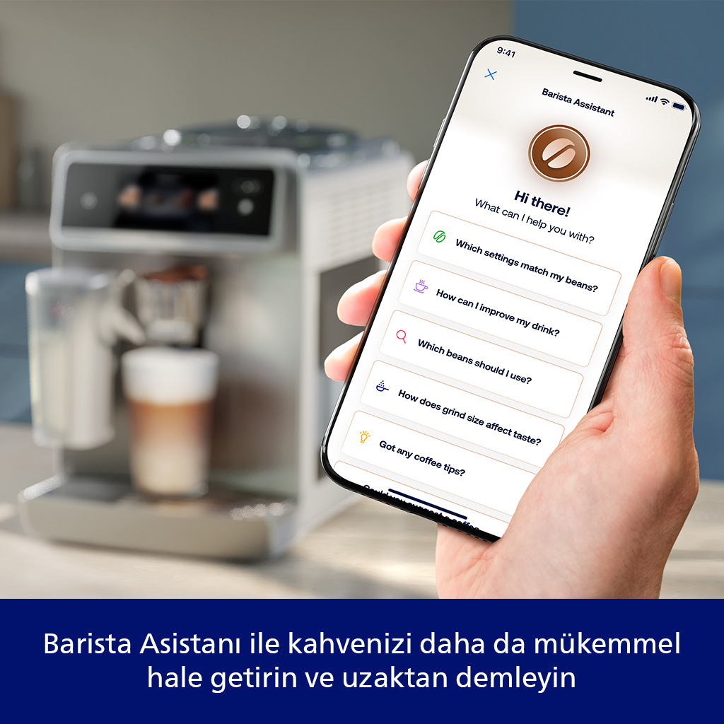 Bir el, bir kahve asistanı uygulaması gösteren bir telefon tutuyor. Arka planda bir kahve makinesi, bir fincan kahve ile. Metin: 'Merhaba!'