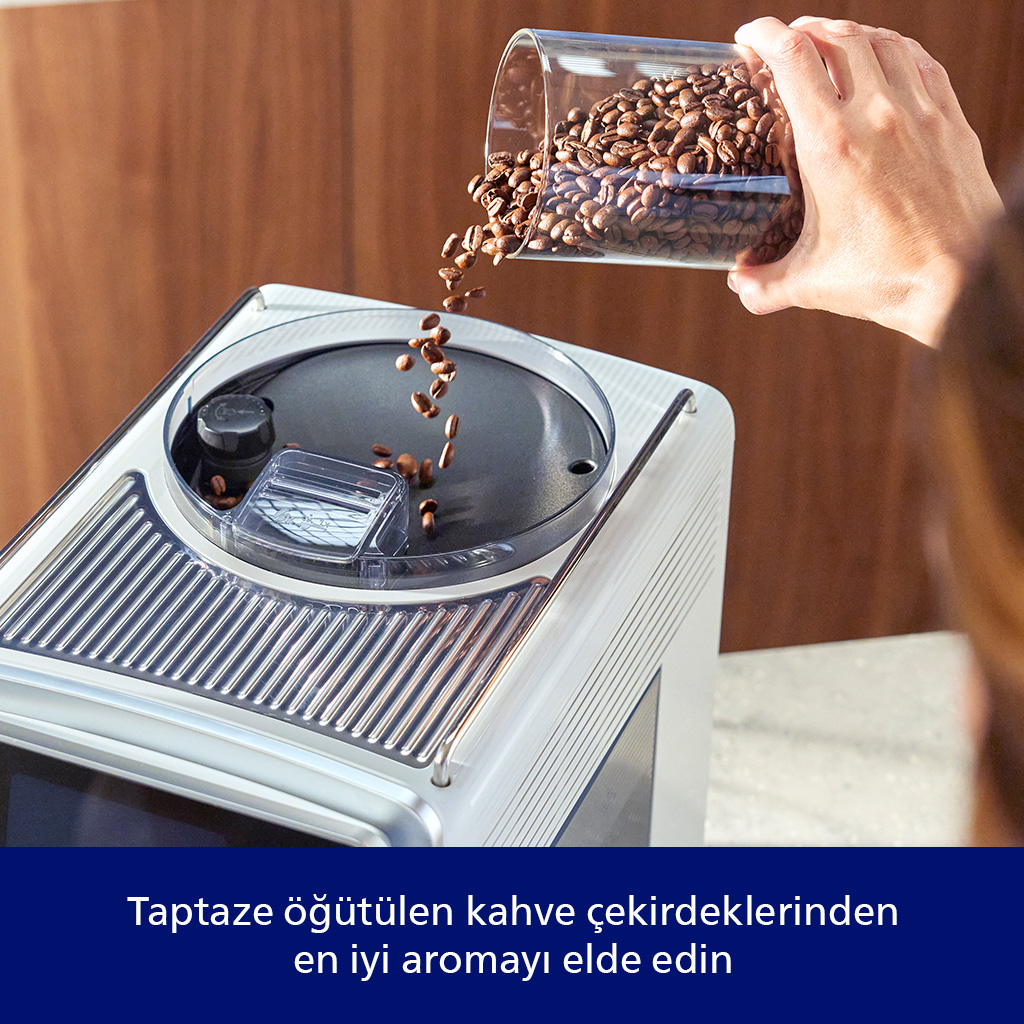 Kahve çekirdekleri kahve makinesine dökülüyor. Bir el, fasulyelerle dolu bir cam kap tutuyor. Makine beyaz ve gümüş renginde.