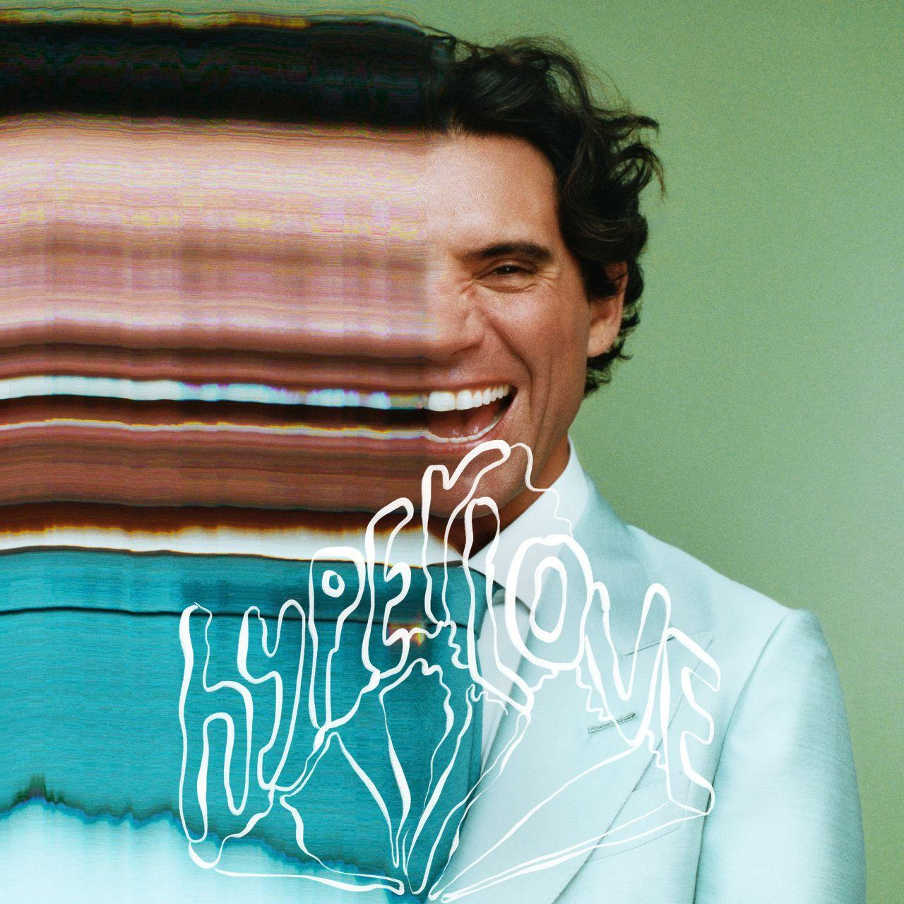 Een man glimlacht breed, draagt een pak. De afbeelding heeft een glitch-effect aan de linkerkant. Het woord 'HYPERLOVE' is overgelegd.