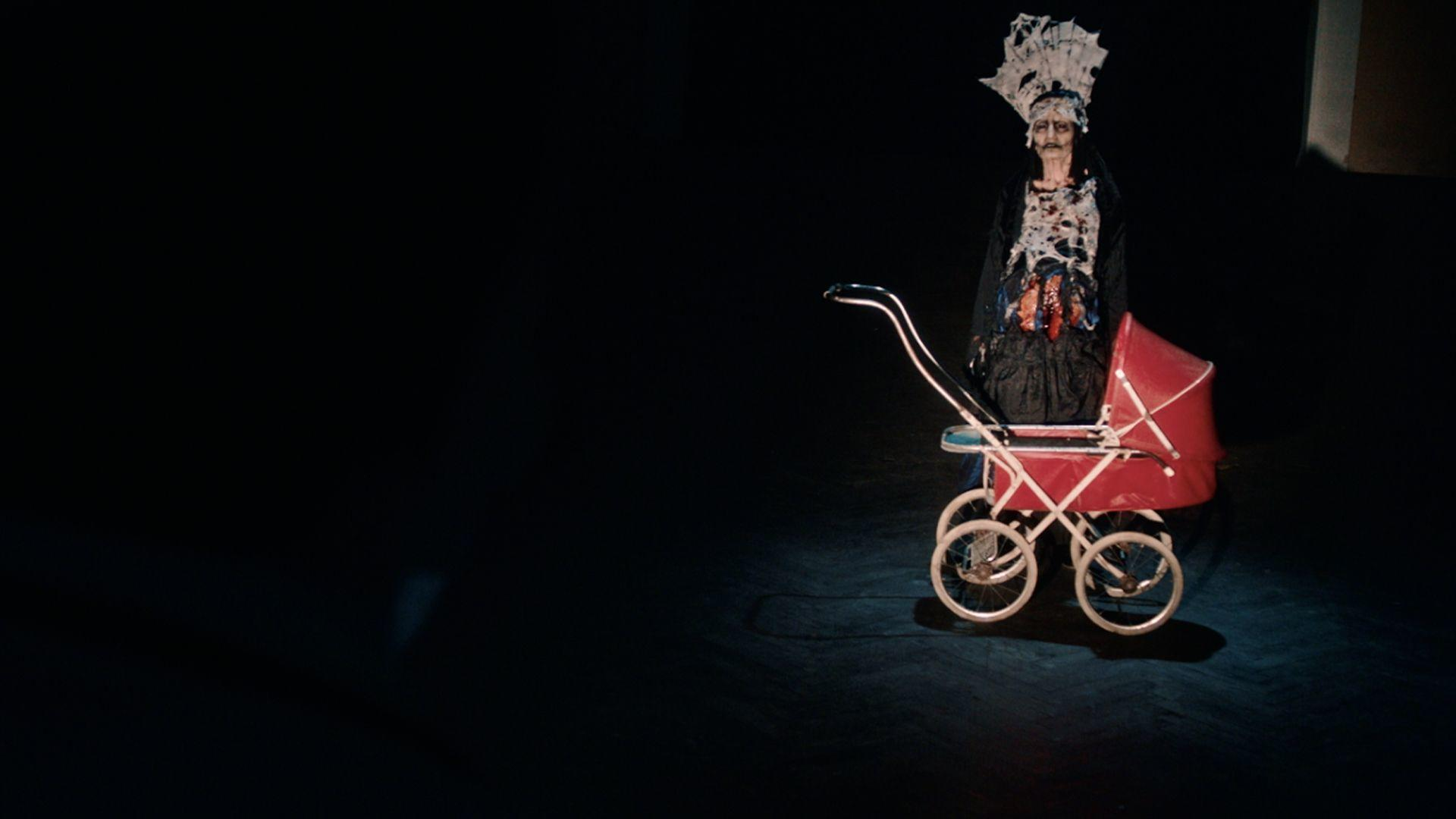 Eine Person in Kostüm steht neben einem roten Kinderwagen. Die Szene ist dunkel, die Person wird beleuchtet. Das Kostüm ist aufwendig und theatralisch.