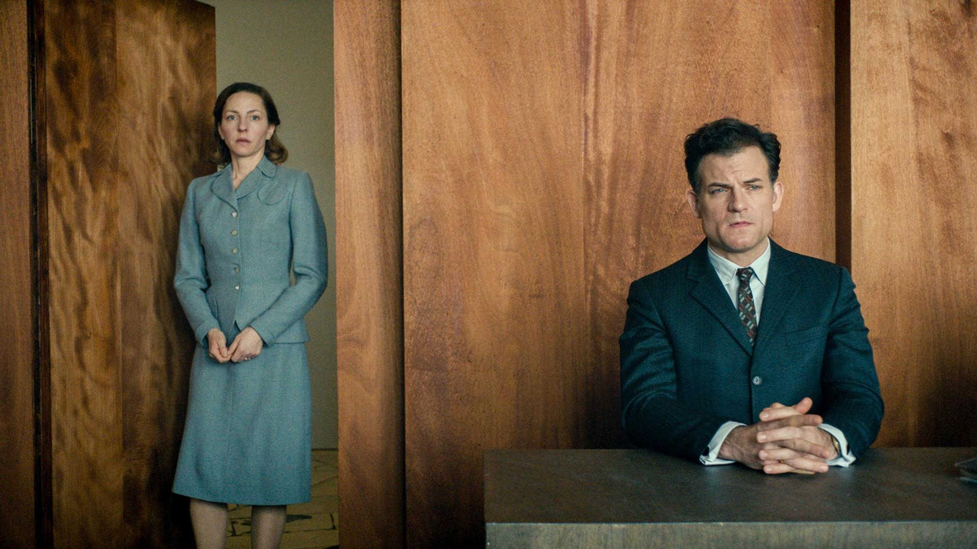 Eine Frau in hellblauem Anzug steht, ein Mann in dunklem Anzug sitzt an einem Tisch. Holzpaneele im Hintergrund.