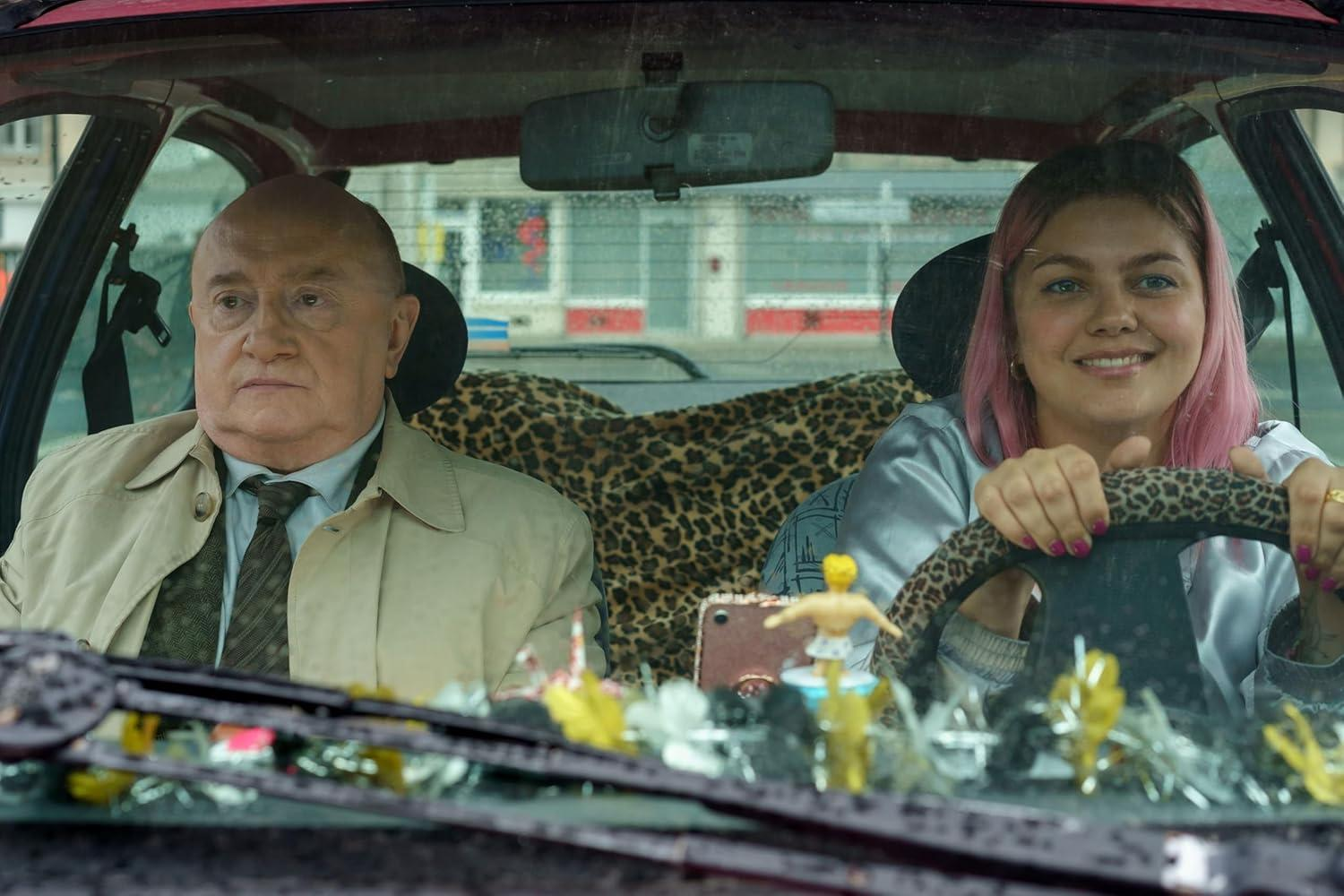 Im Auto sind zwei Personen anwesend. Ein kahler Mann links und eine Frau mit rosa Haaren rechts. Die Frau lächelt.