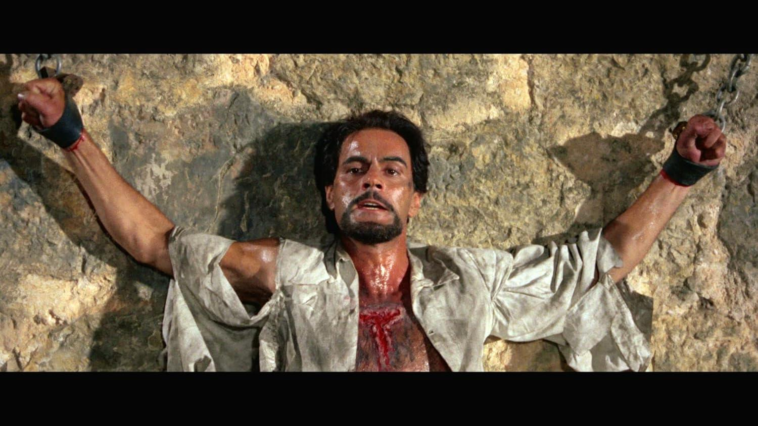 Mann mit Bart, Arme an Felsenwand gefesselt, zerrissenes Hemd, Brustwunde, Blut. Historische Kulisse.