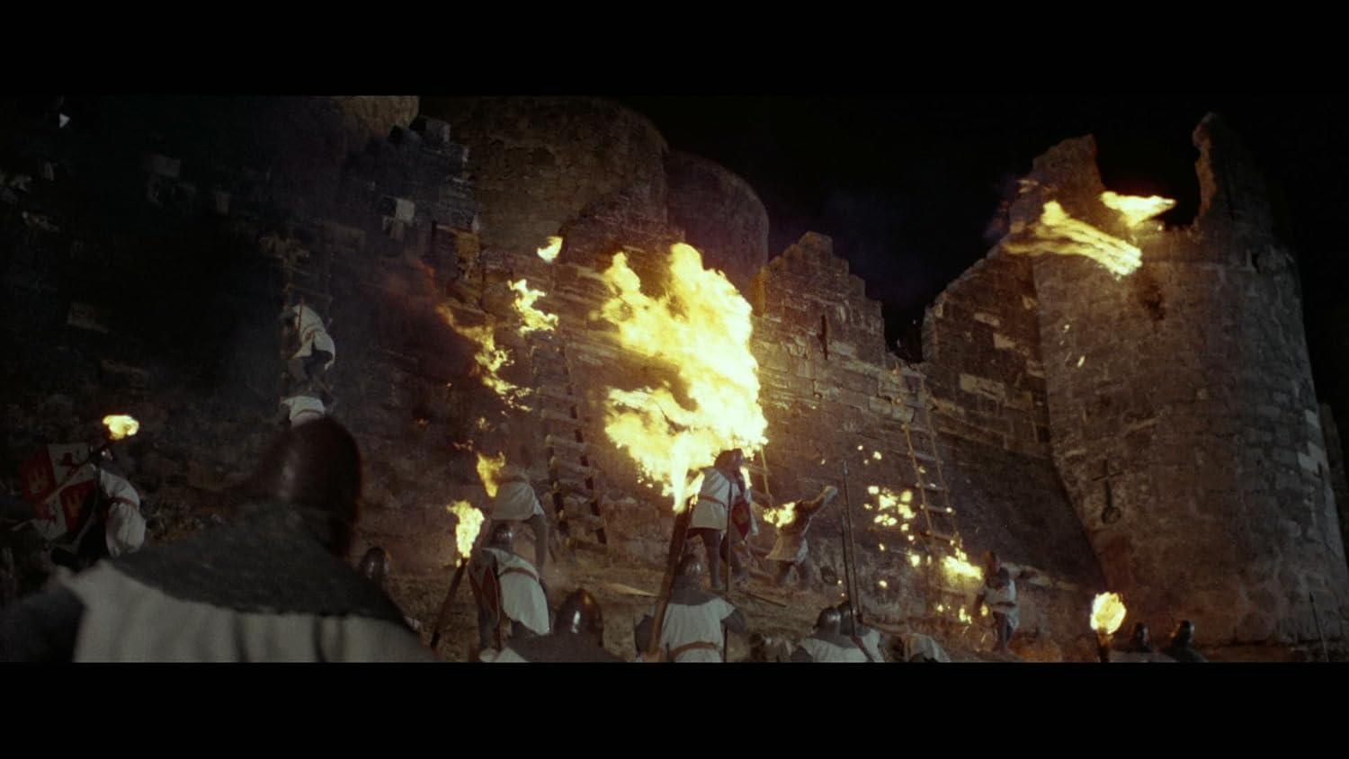 Mittelalterliche Burg unter Belagerung bei Nacht. Flammen verschlingen die Mauer. Soldaten in weißen Tuniken klettern Leitern hinauf.