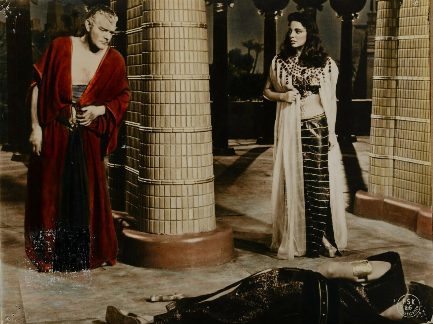 Szene aus einem Film. Ein Mann und eine Frau stehen in der Nähe einer Säule. Eine Person liegt am Boden, möglicherweise tot.