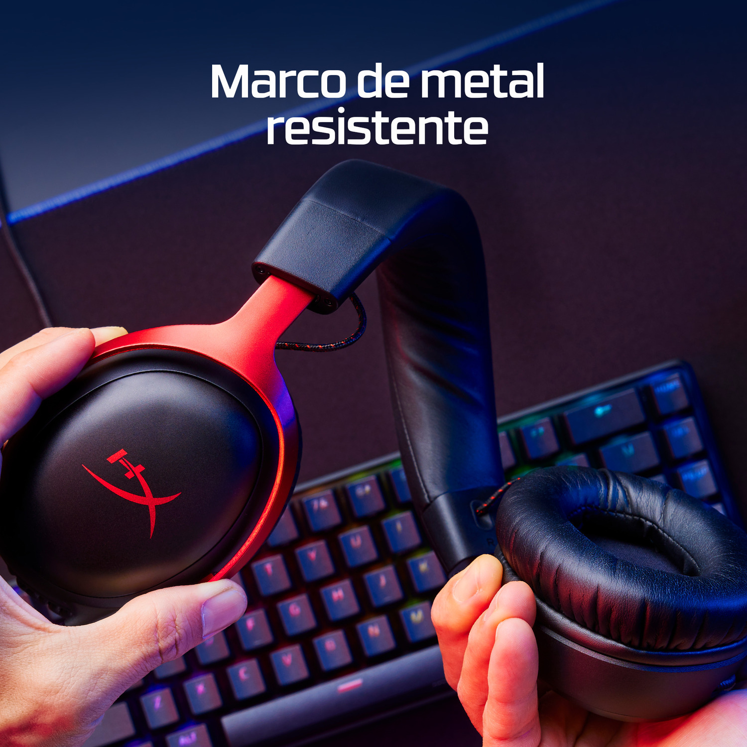 Manos sosteniendo unos auriculares gaming negros y rojos, con un teclado en el fondo. Los auriculares tienen el logo de HyperX.