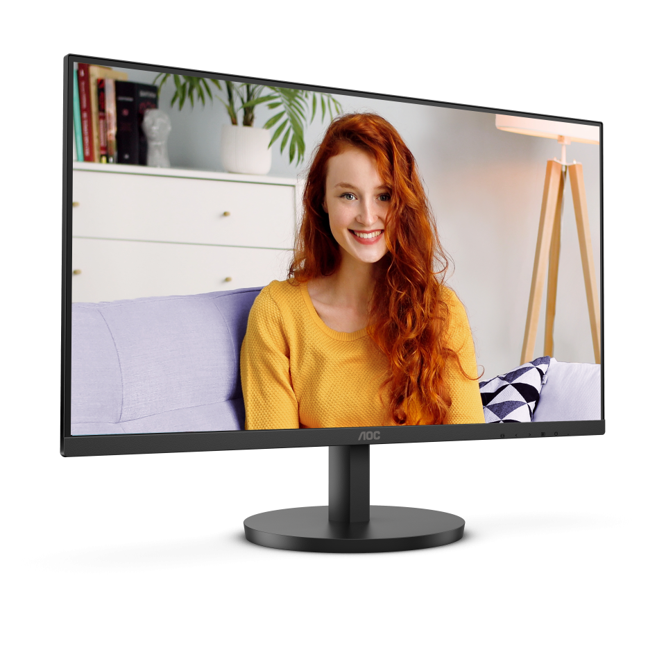 Egy számítógép monitor egy vörös hajú, sárga pulóveres nőt mutat. A háttérben egy szoba látható bútorokkal és egy növénnyel.