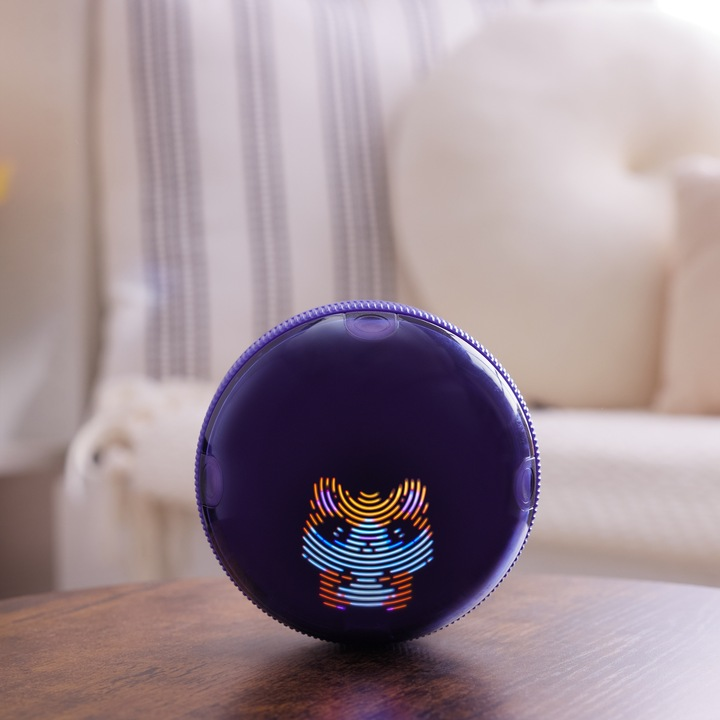 Fioletowe, okrągłe urządzenie z grafiką kota na drewnianym stole. Tło przedstawia krzesło, poduszki i okno.