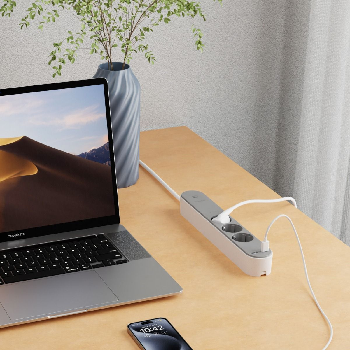 Ein Schreibtisch mit Laptop, Telefon, Steckdosenleiste und Vase mit Pflanzen. Im Hintergrund eine Wand und ein Vorhang.