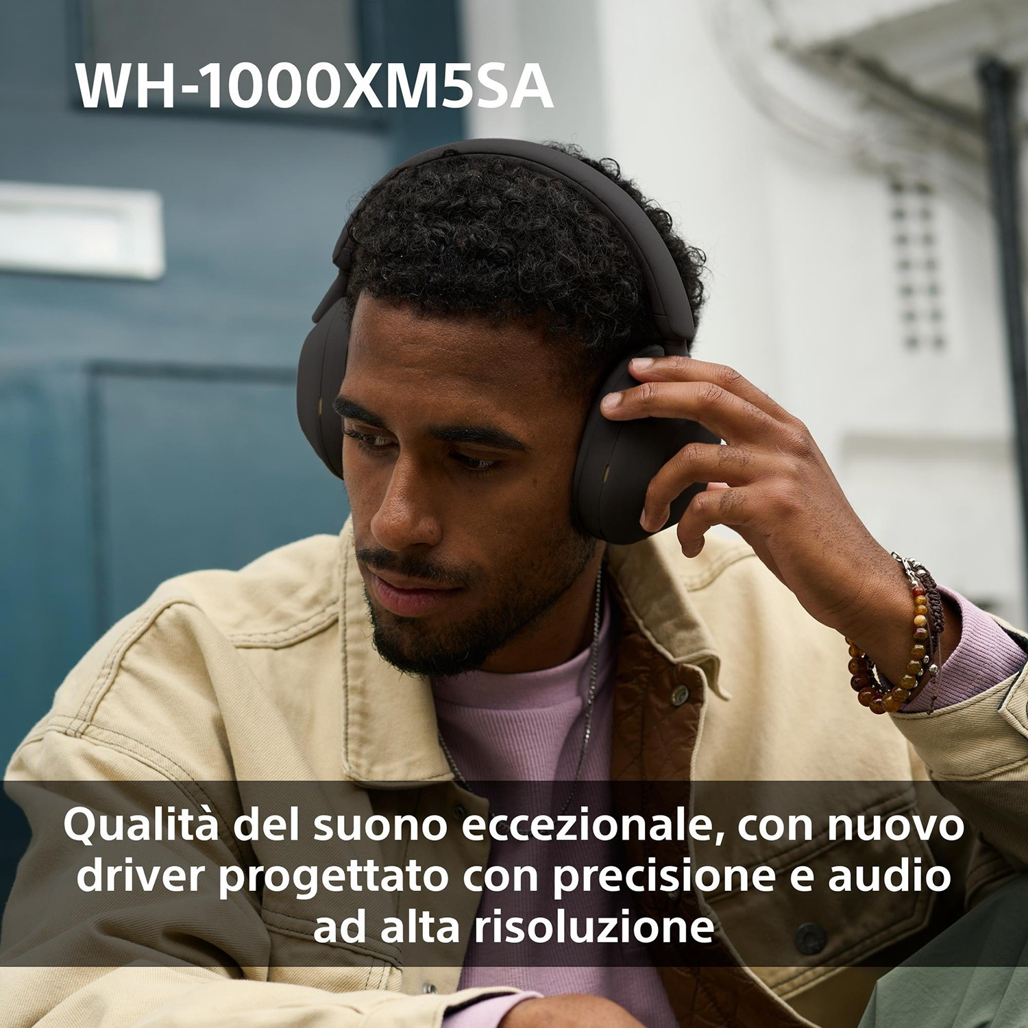 Un uomo con cuffie nere le tocca con la mano destra. Indossa una giacca beige e una camicia rosa. Testo in basso.