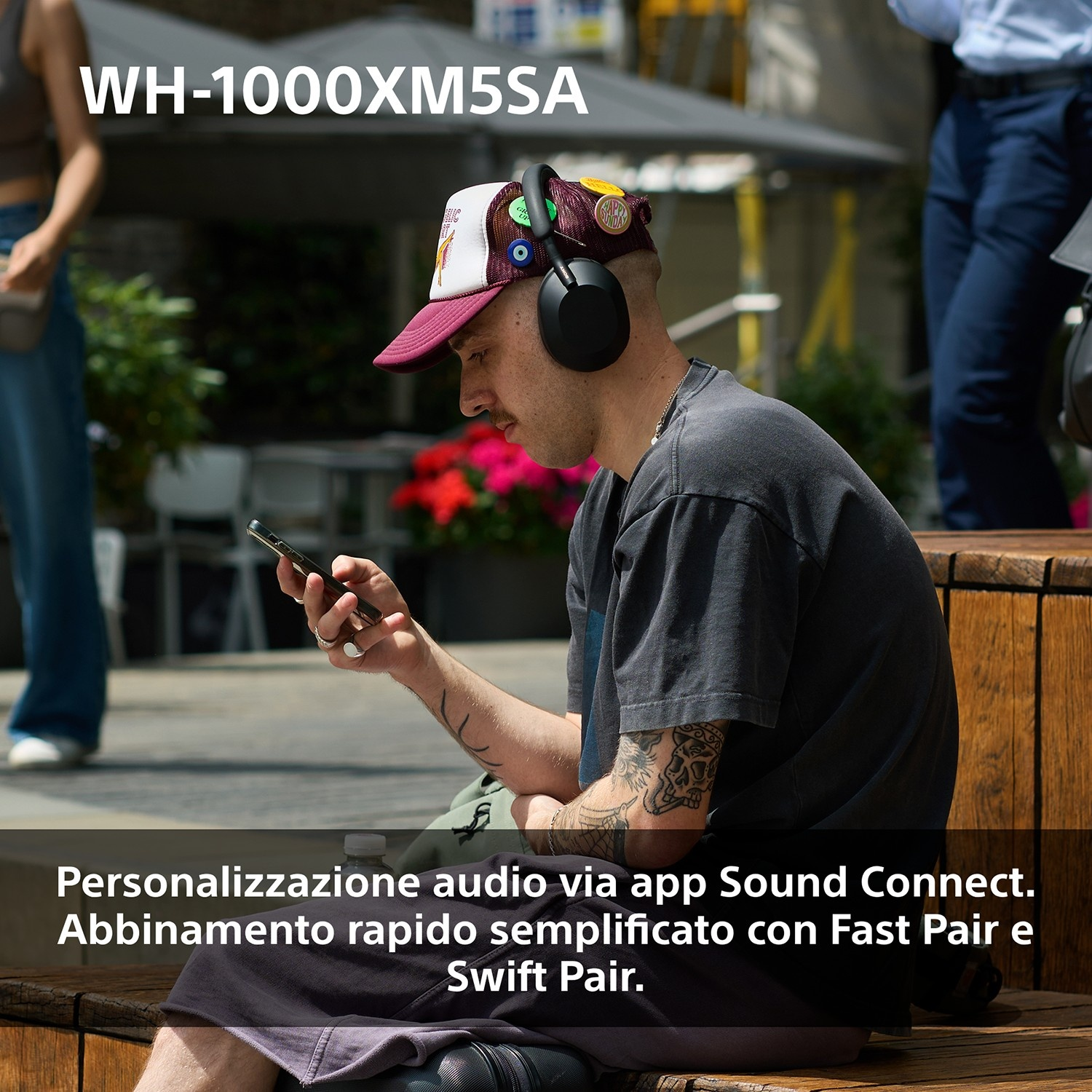 Un uomo con le cuffie e un berretto guarda il suo telefono. Lo sfondo include persone e il testo 'WH-1000XM5SA'.