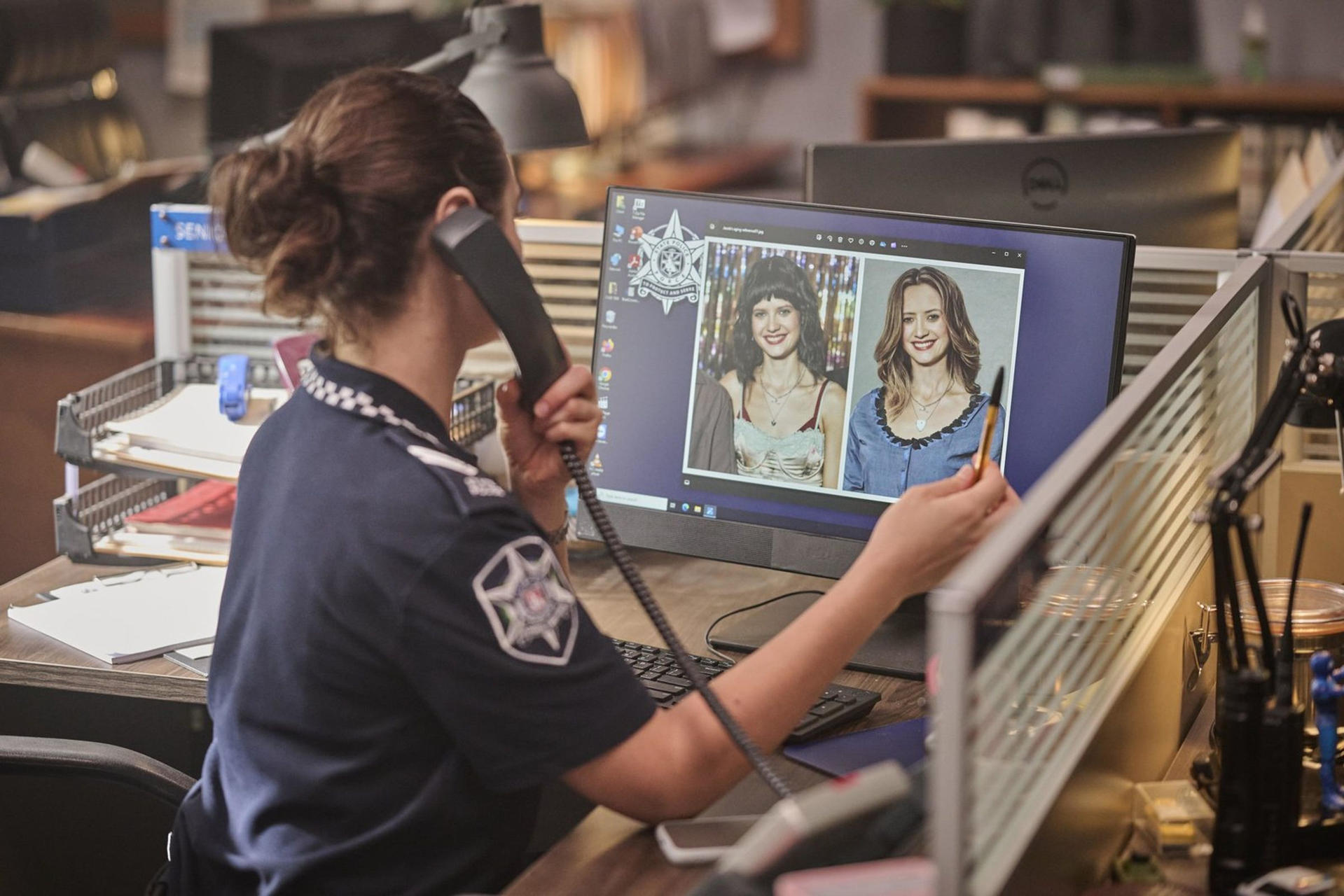 Eine Polizistin telefoniert und betrachtet Fotos von zwei Frauen auf einem Computerbildschirm. Büroumgebung mit Schreibtischen und Bildschirmen.