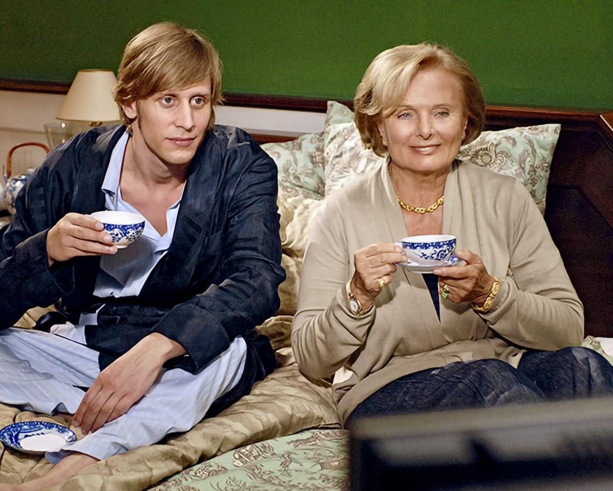 Zwei Personen trinken Tee auf einem Bett. Ein Mann trägt einen Bademantel, die Frau einen Pullover. Beide halten Teetassen mit blauem Muster.