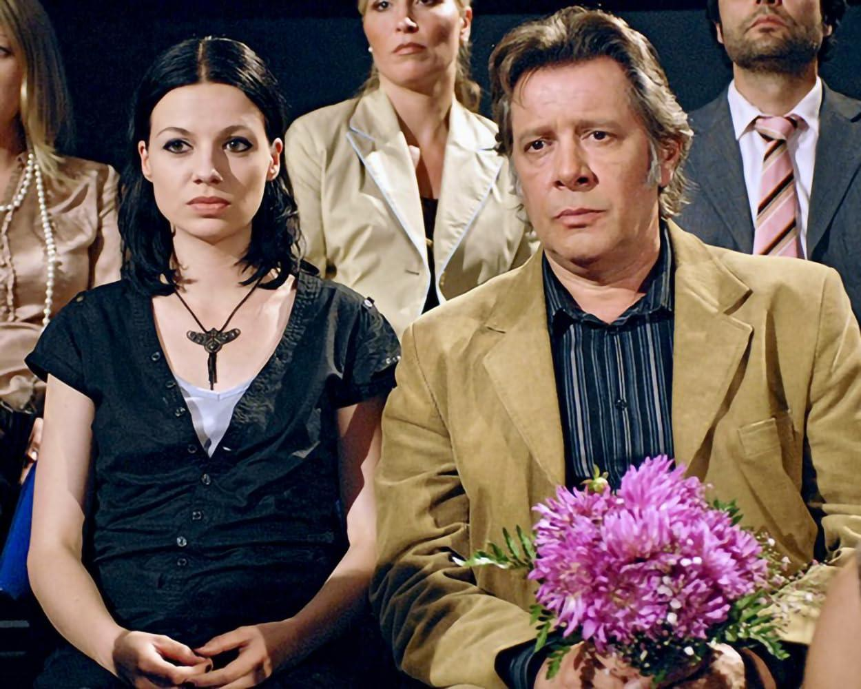 Ein Mann hält rosa Blumen, neben einer Frau mit Halskette. Andere sitzen im Hintergrund. Formelle Kleidung ist zu sehen.