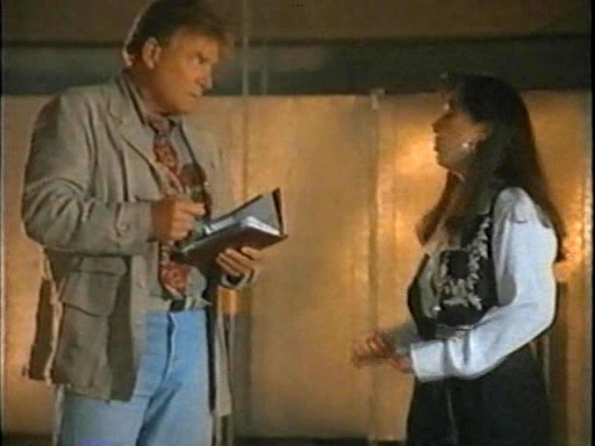 Ein Mann in Jacke und Jeans hält ein Buch und blickt eine Frau im Kleid und weißer Bluse an. Beide sind drinnen.