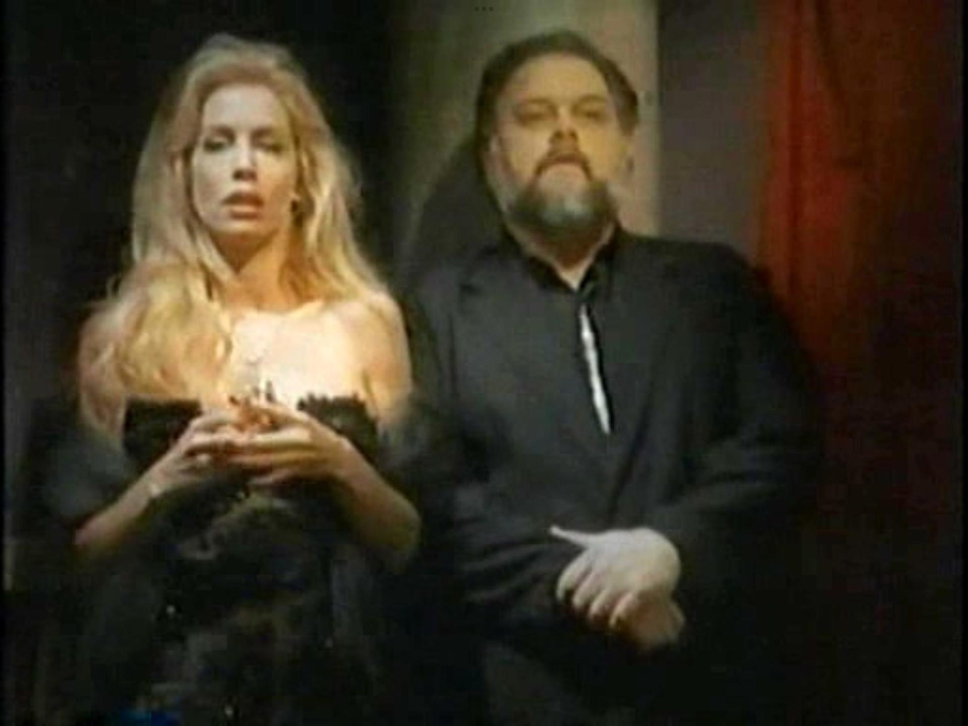 Ein Mann und eine Frau stehen eng beieinander. Die Frau hat lange blonde Haare. Der Mann hat einen Bart und trägt einen dunklen Anzug.