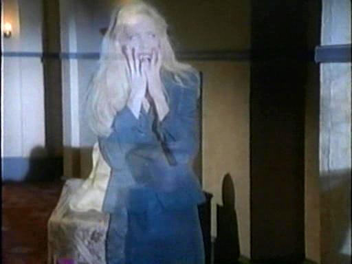Eine Frau mit blonden Haaren in einem blauen Anzug bedeckt ihr Gesicht mit den Händen und sieht verängstigt aus. Sie ist in einem Raum.