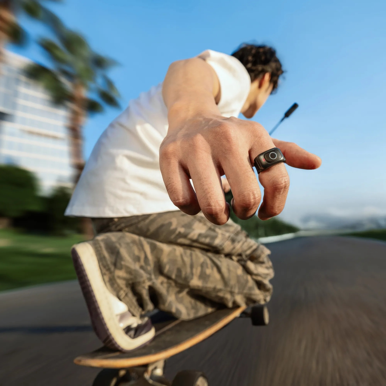 Una persona su uno skateboard, con un anello con schermo. La persona indossa una camicia bianca e pantaloni mimetici. Cielo blu sullo sfondo.