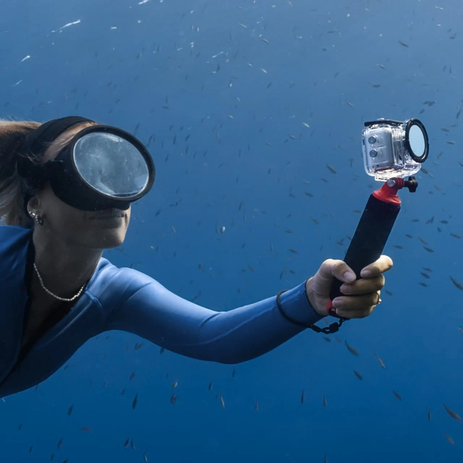 Un subacqueo in attrezzatura blu sott'acqua tiene una fotocamera. Numerosi piccoli pesci nuotano intorno. Il subacqueo indossa una maschera.