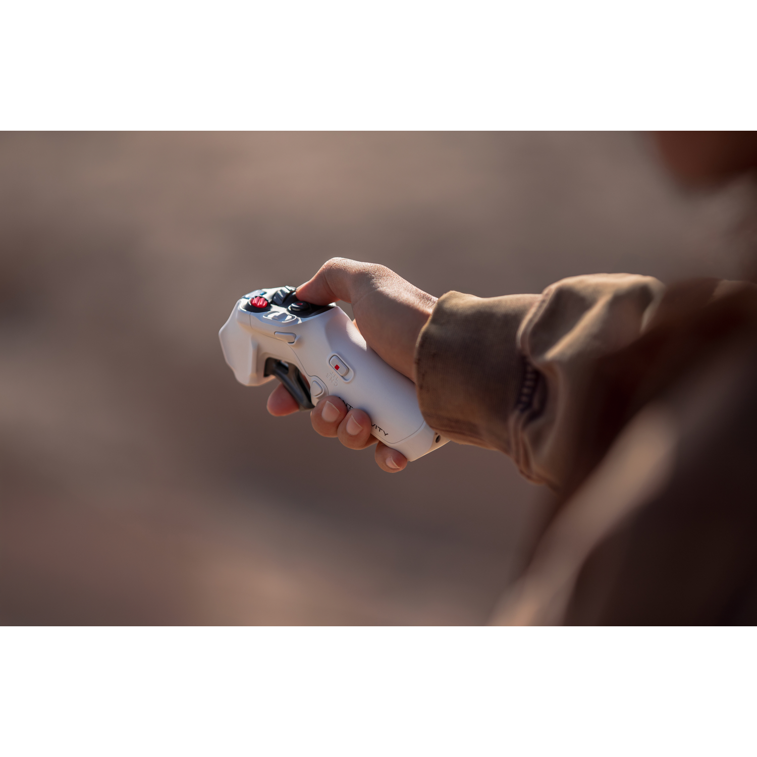 Een persoon houdt een witte controller met rode knoppen vast. De achtergrond is wazig en toont een bruine buitenomgeving.