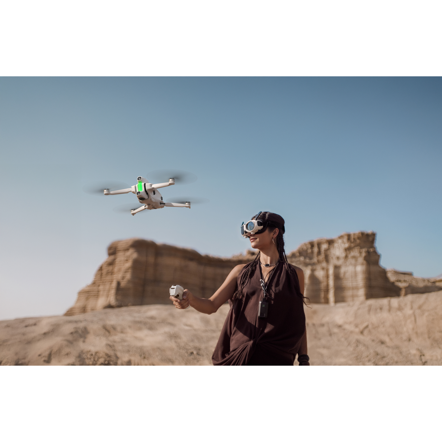 Een vrouw met een bril en een controller bestuurt een drone in een woestijnlandschap met een heldere blauwe lucht.