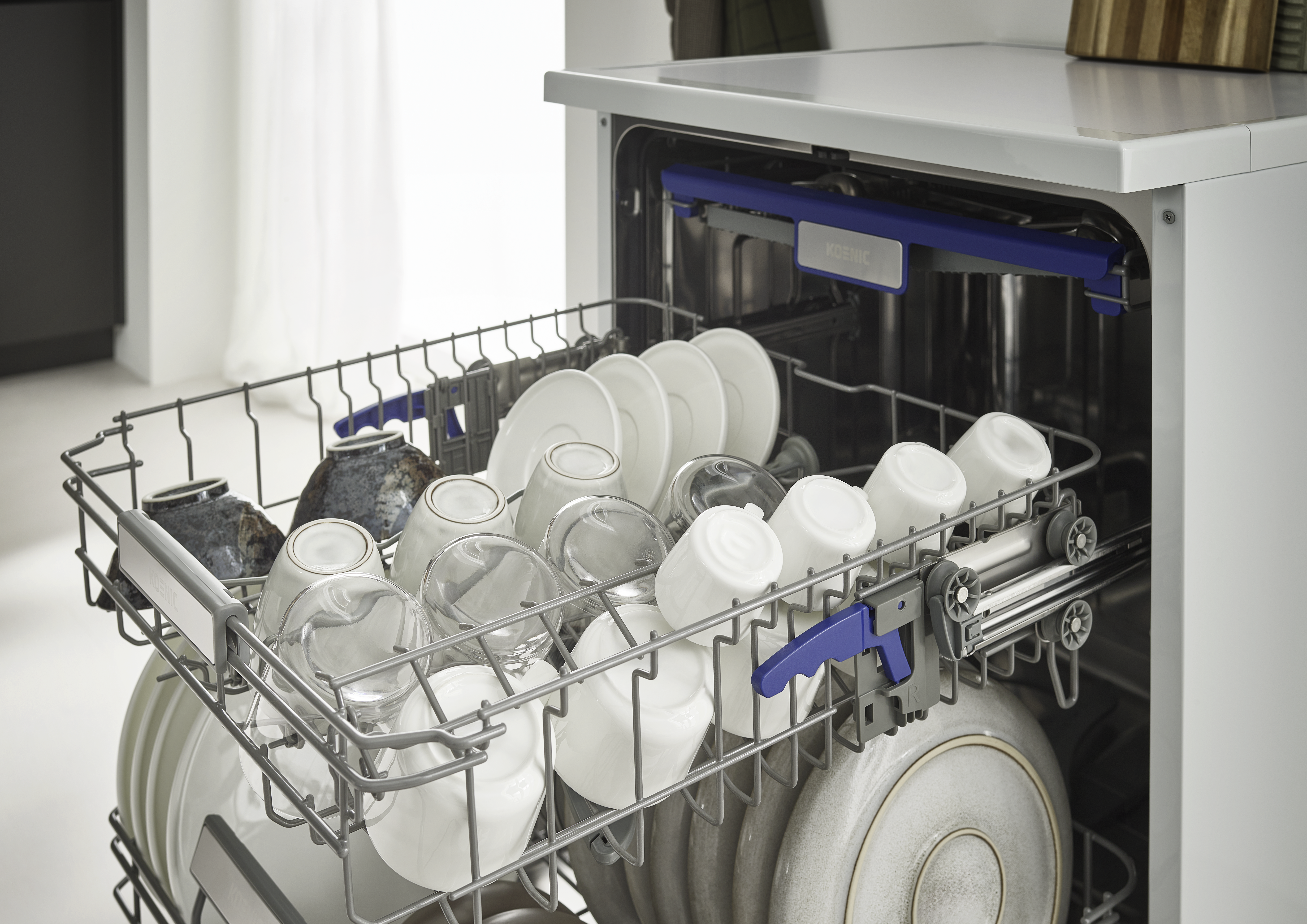 Geschirrspüler mit offener Tür, beladen mit Geschirr, Tassen und Gläsern. Weiße und graue Töne dominieren die Szene.