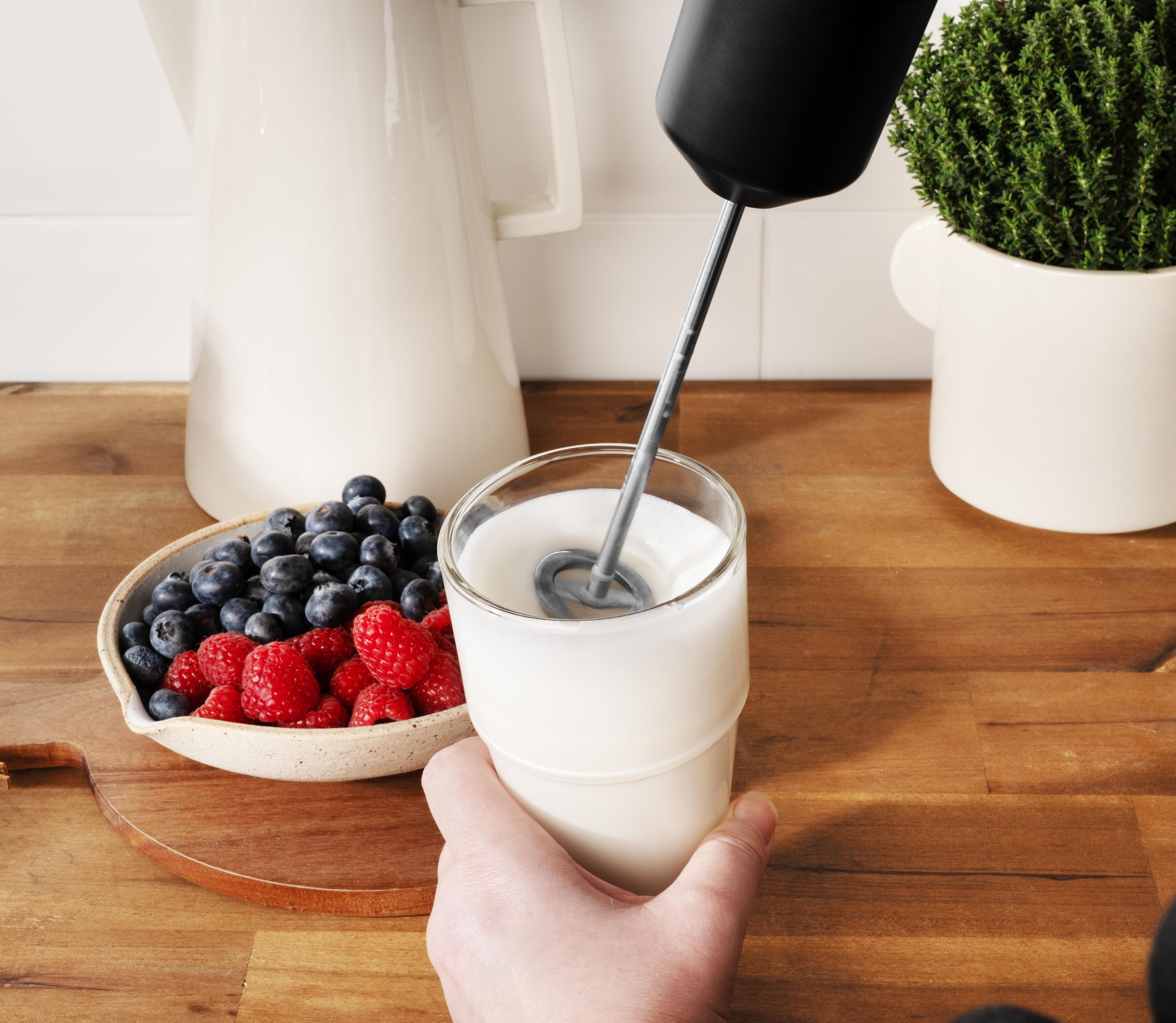 Eine Hand hält ein Glas mit aufgeschäumter Milch. Blaubeeren und Himbeeren sind in einer Schüssel daneben.