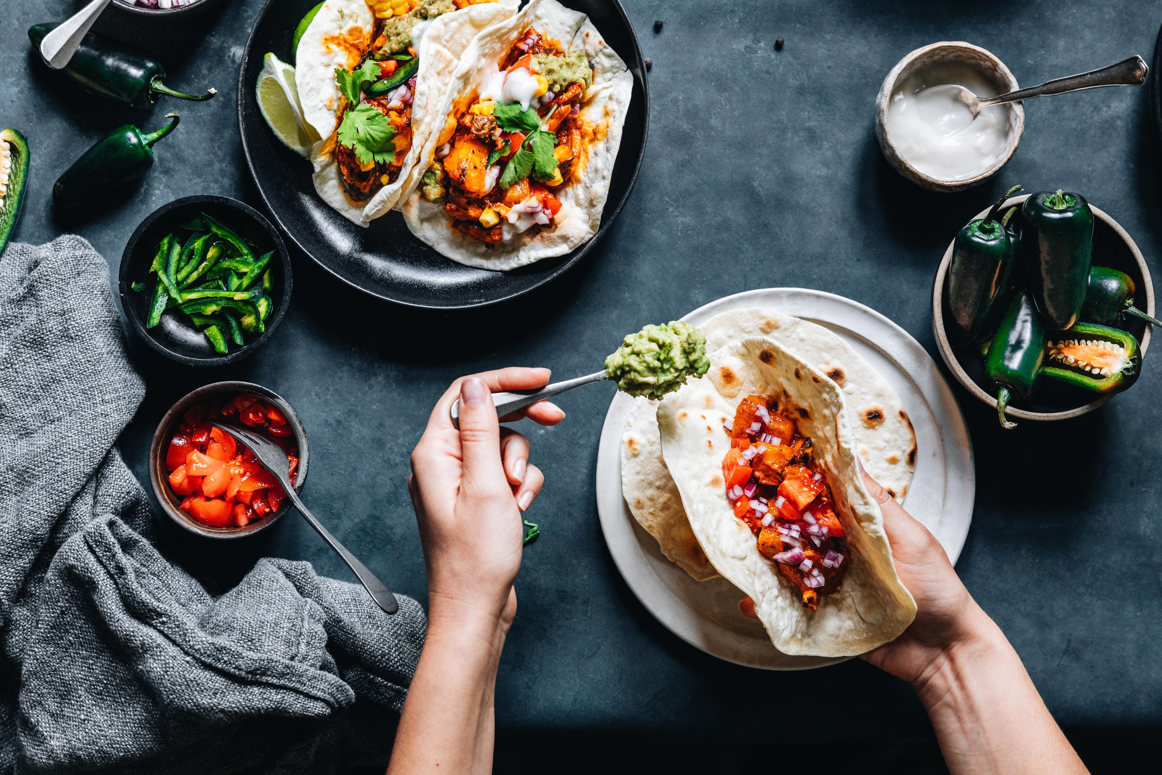 Draufsicht auf Tacos mit Belag, Hände fügen Guacamole hinzu. Zutaten sind Paprika, Tomaten und Saucen. Dunkle Oberfläche.