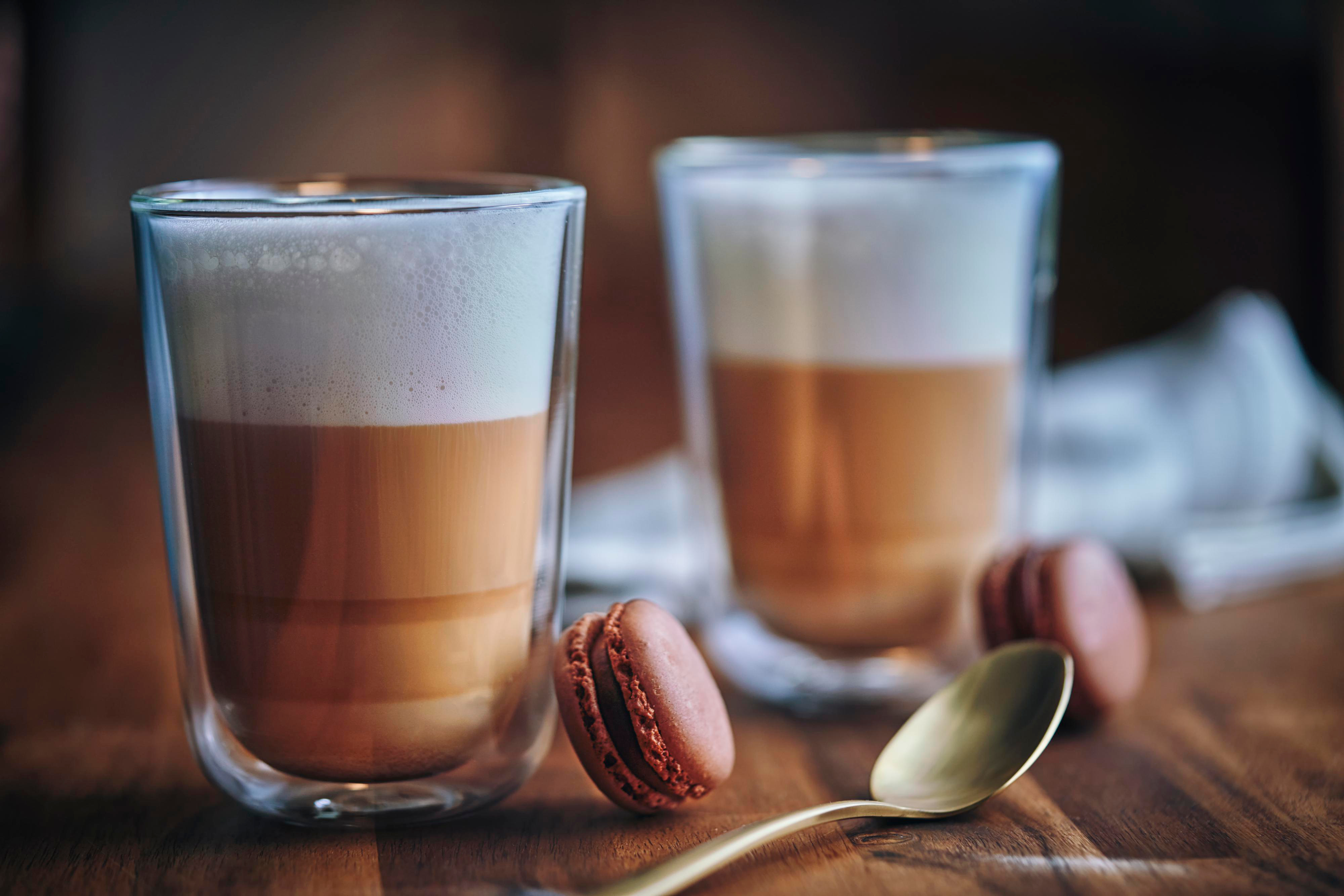 Zwei Lattes in Glasbechern, Makronen und ein Löffel auf einer Holzoberfläche.