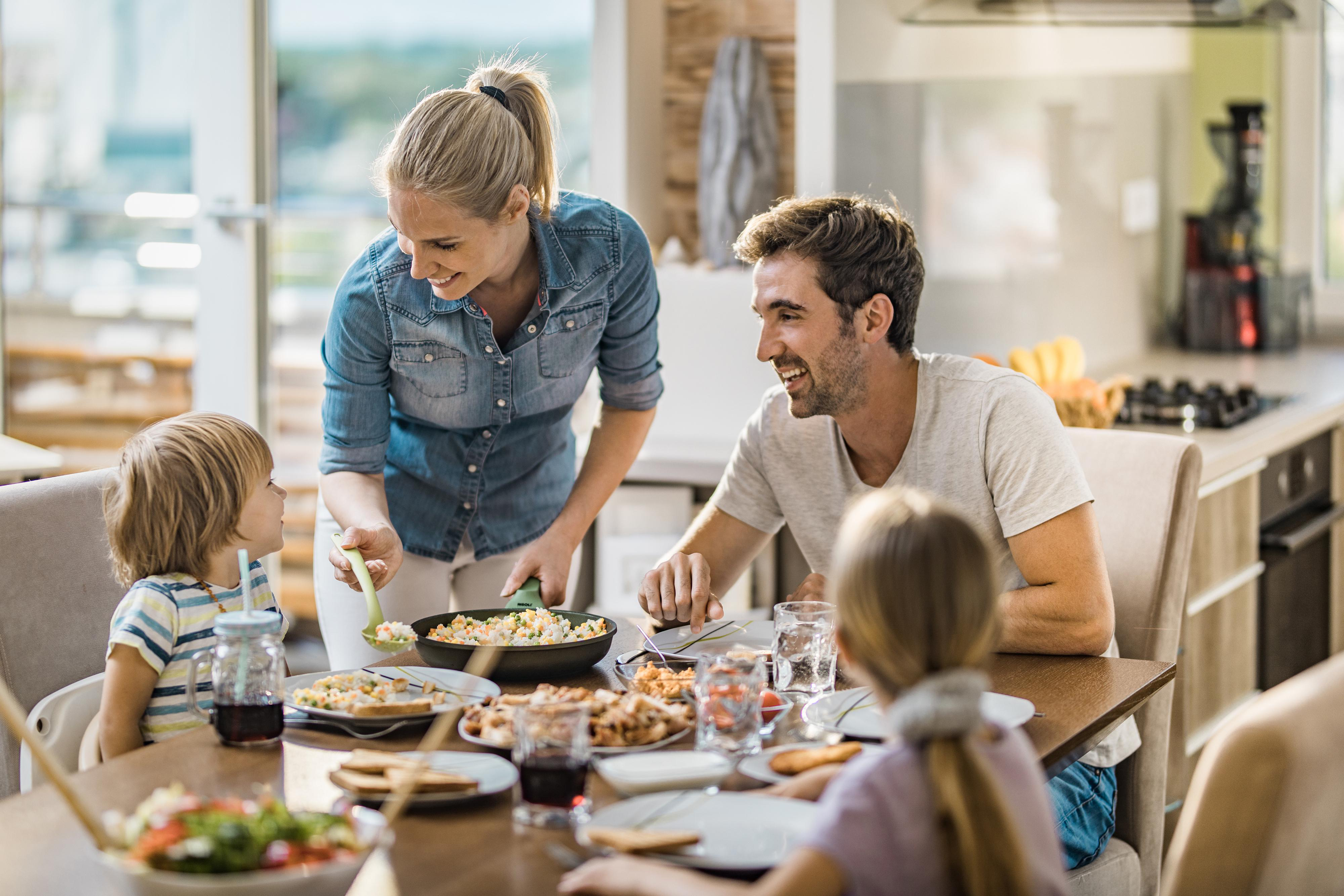 Familie am Tisch. Eine Frau serviert Essen aus einer Pfanne. Andere sitzen und lächeln. Essen und Getränke stehen auf dem Tisch.