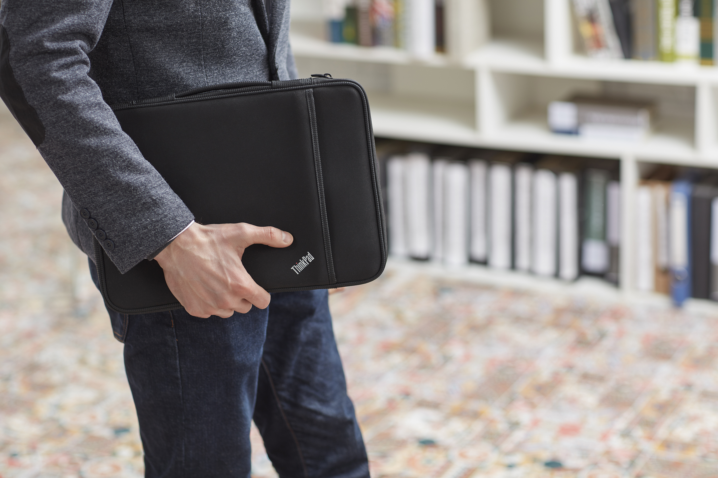 Egy személy egy fekete ThinkPad laptop tokot tart, szürke zakót és farmert visel, egy könyvespolc közelében állva.