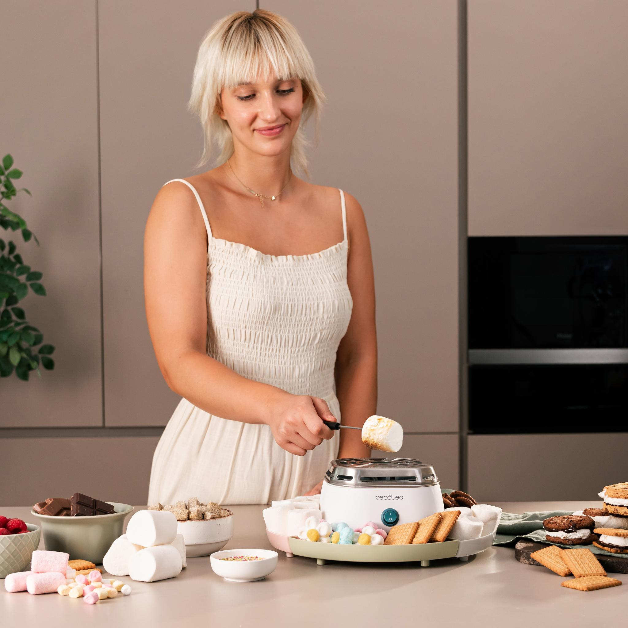 Mujer asando malvavisco sobre un fabricante de s'mores. Se ven ingredientes como chocolate, galletas y malvaviscos.