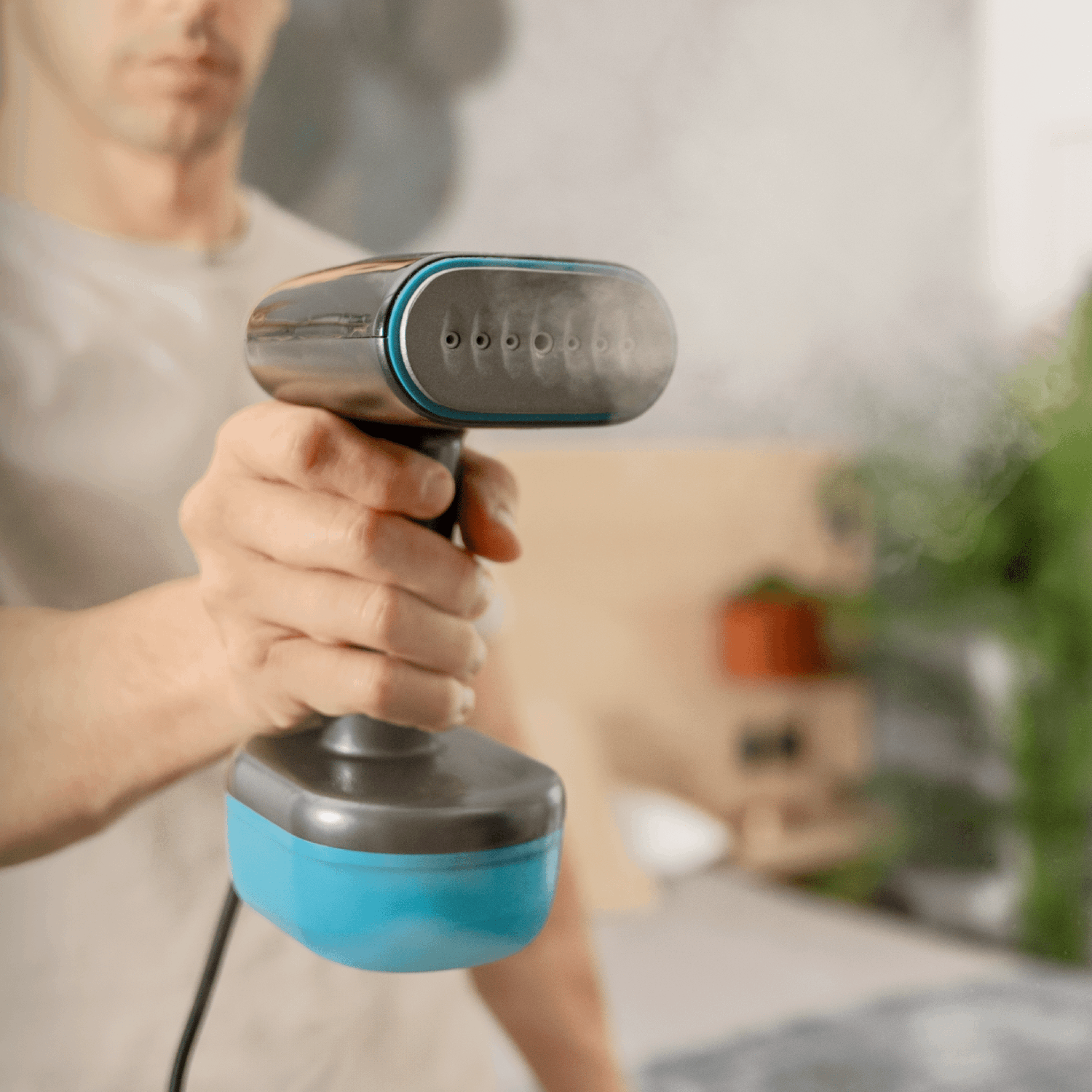 Una persona sostiene un vaporizador de ropa azul y negro, emitiendo vapor. El fondo incluye una planta y una estantería de madera.