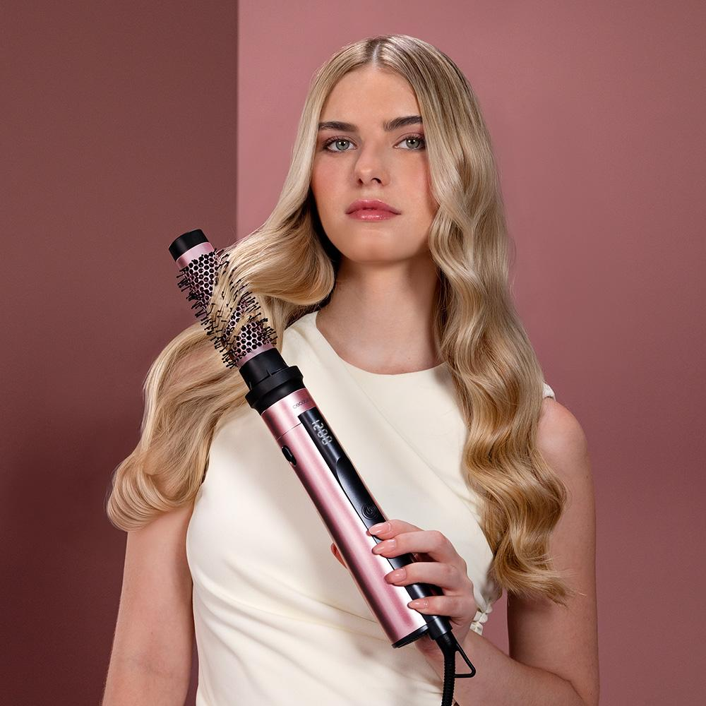 Mujer rubia sosteniendo un cepillo rosa para peinar el cabello. Lleva una camisa blanca con fondo rosa. Pelo largo y ondulado.