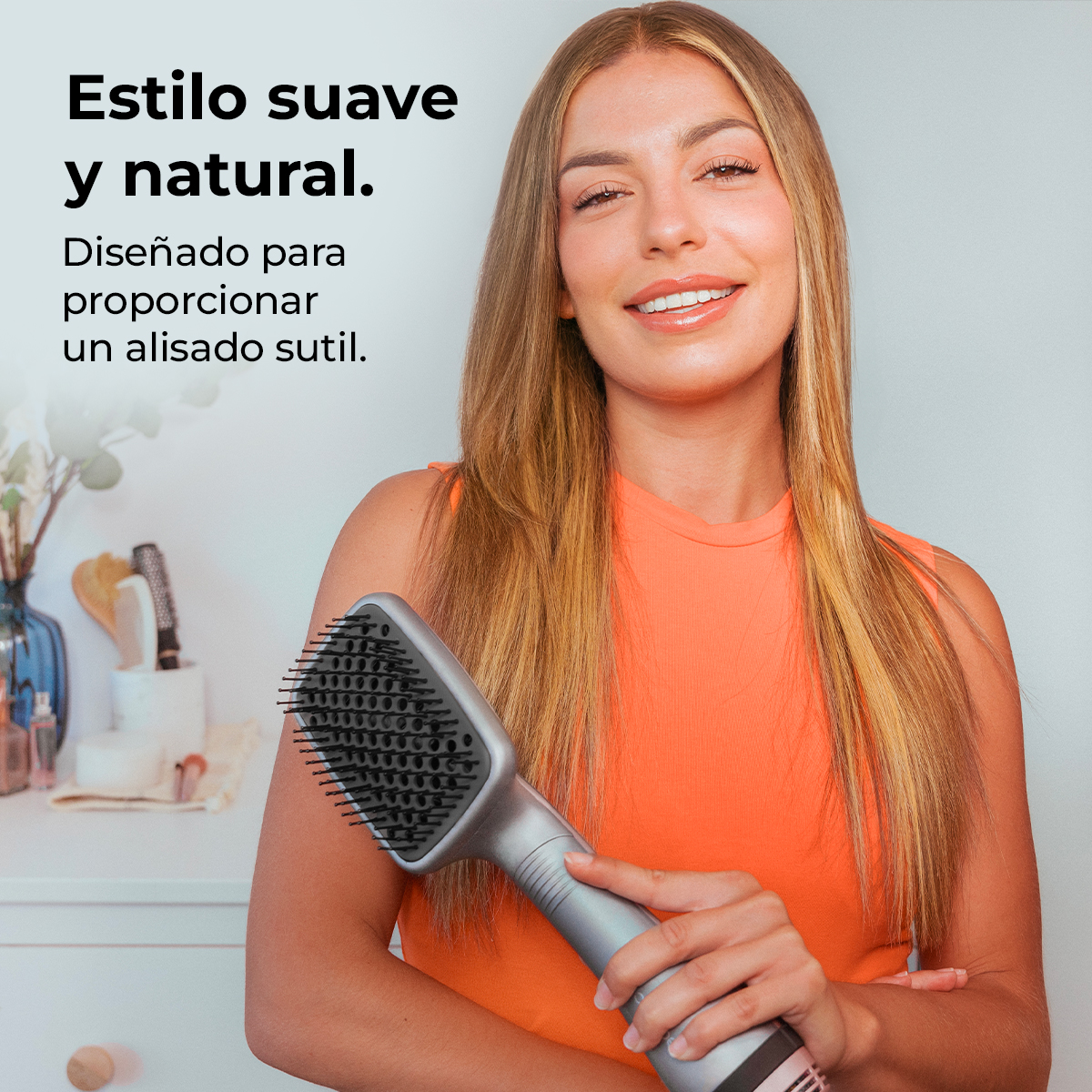 Una mujer sostiene una herramienta gris para peinar el cabello. Lleva una blusa naranja y sonríe. El texto en español dice 'Estilo suave y natural'.