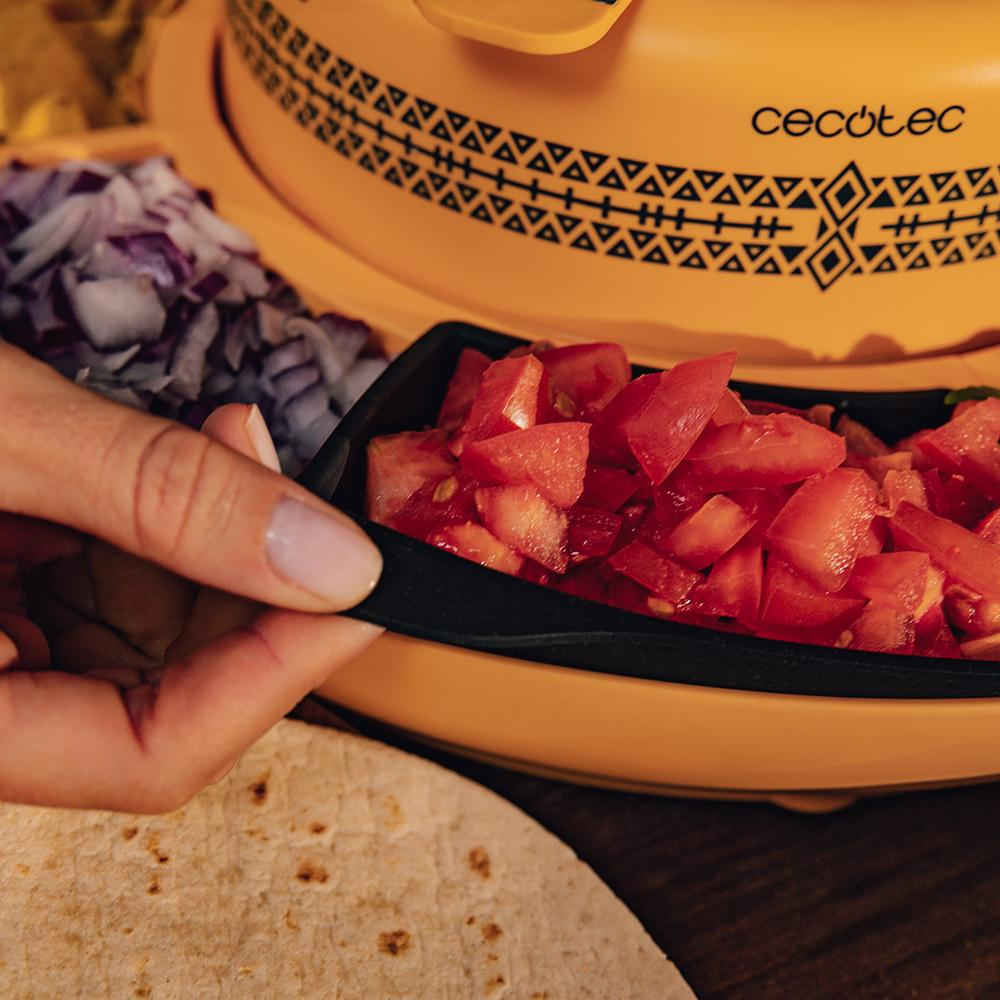 Mano sosteniendo una bandeja negra con tomates picados. Cocina amarilla y tortilla en primer plano. Cebollas rojas y nachos en el fondo.