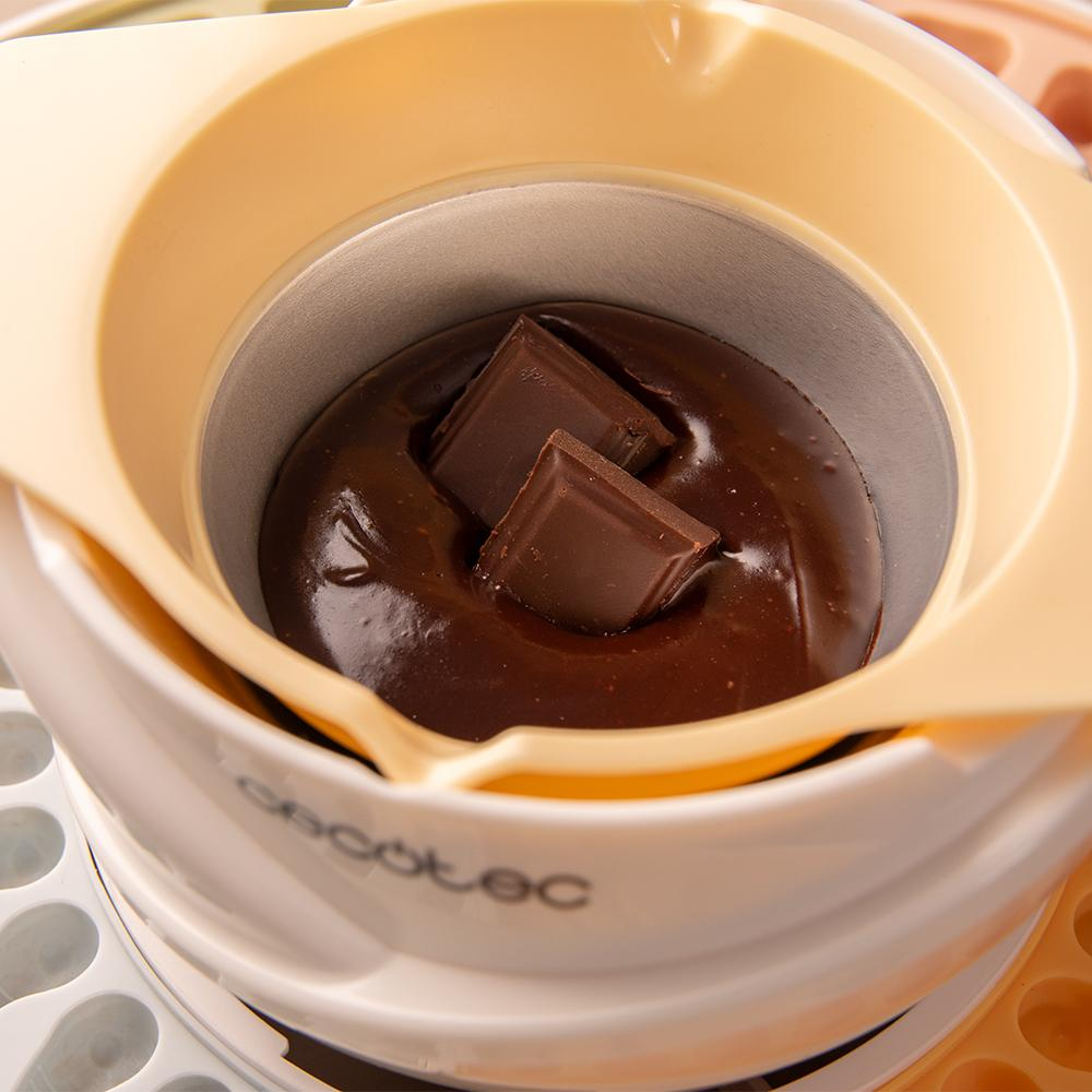 Primer plano de una olla para derretir chocolate. Dos cuadrados de chocolate se encuentran en chocolate derretido. La olla está dentro de un soporte color crema.