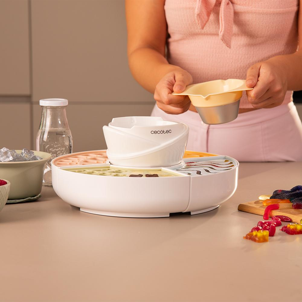 Una persona sostiene un cuenco sobre un aparato de cocina blanco. Ingredientes y una tabla de cortar con caramelos en la mesa.