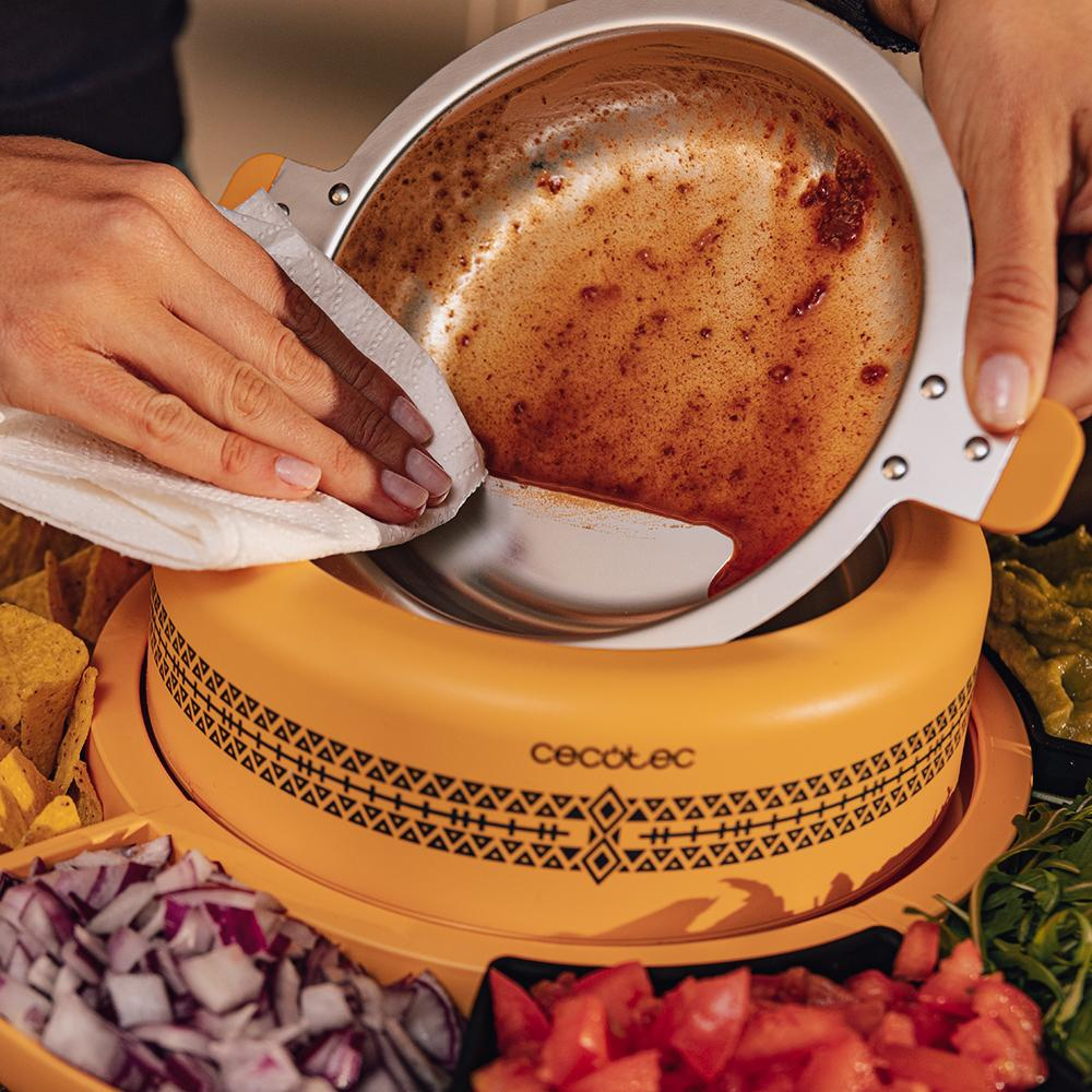 Manos limpiando un cuenco de metal sucio sobre un calentador de alimentos amarillo. Los ingredientes incluyen salsa, guacamole y cebollas.