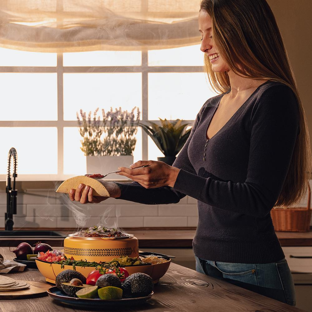 Una mujer sonríe mientras rellena un taco. Vapor se eleva de la comida en una mesa con ingredientes y una cocina en el fondo.
