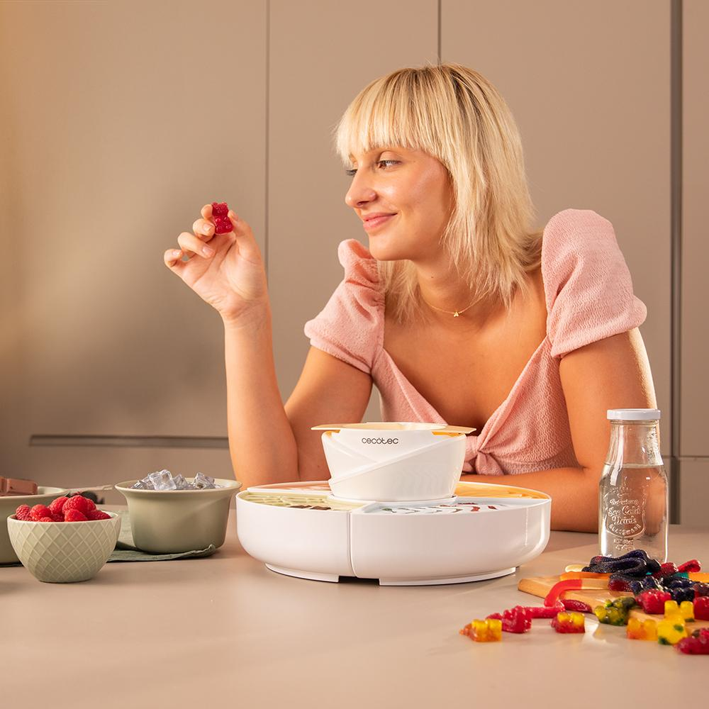 Mujer rubia sosteniendo un osito de goma rojo. Un deshidratador de alimentos, frambuesas y otros ositos de goma están en la mesa.