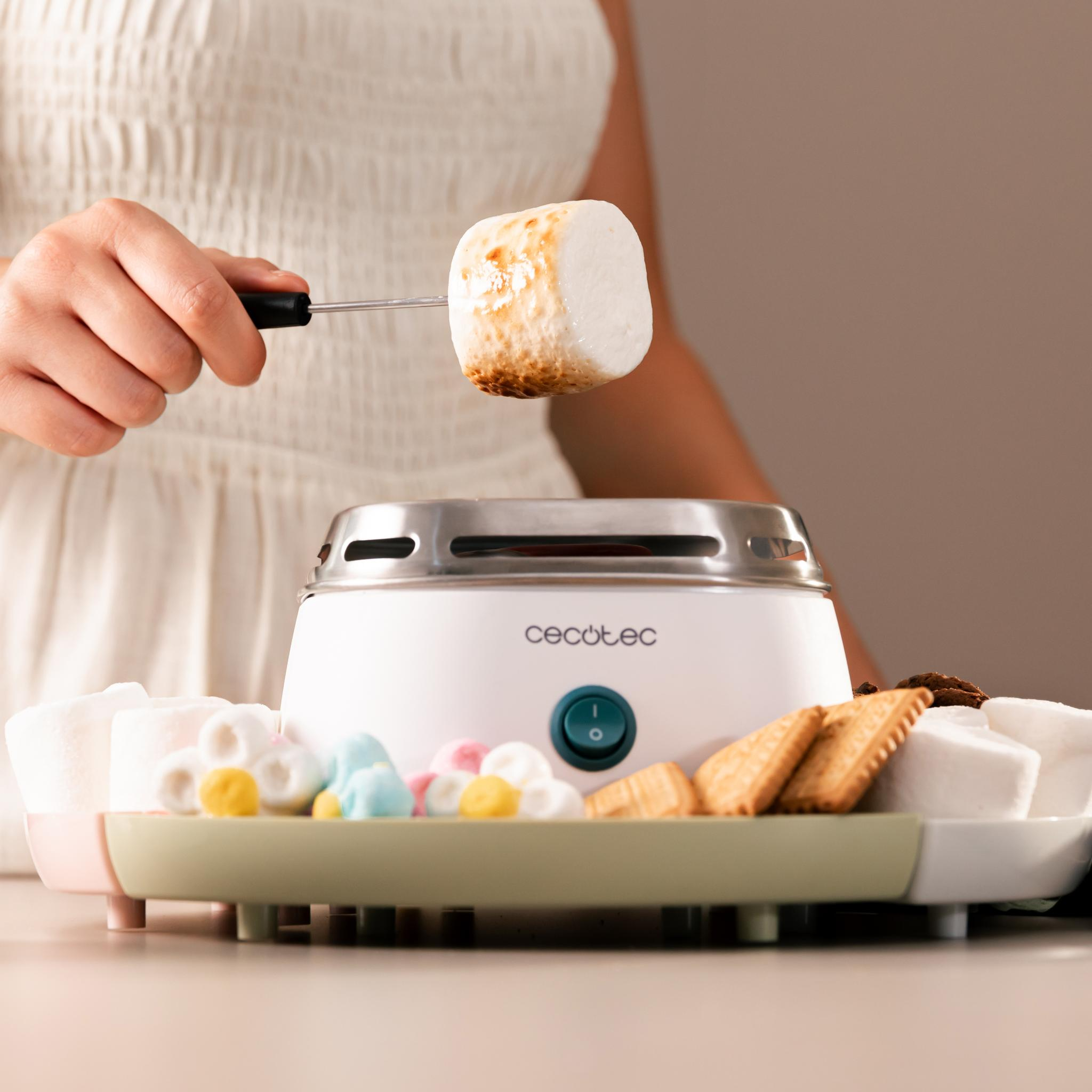 Una persona tuesta una malvavisco. Una máquina blanca para s'mores está en una bandeja con malvaviscos, galletas y dulces.