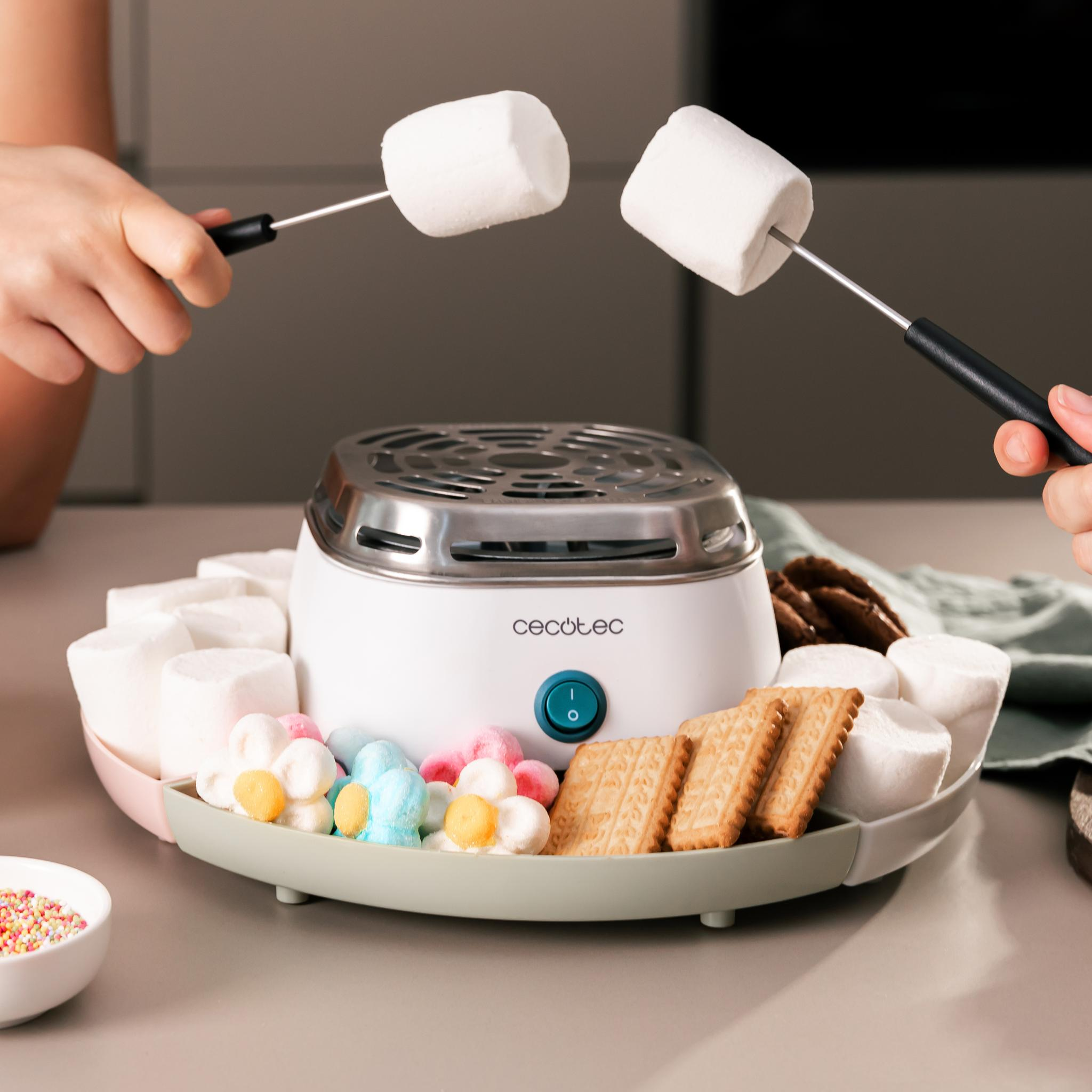 Manos sostienen brochetas con malvaviscos sobre una máquina de s'mores. Galletas, malvaviscos de colores y chispas rodean la máquina.