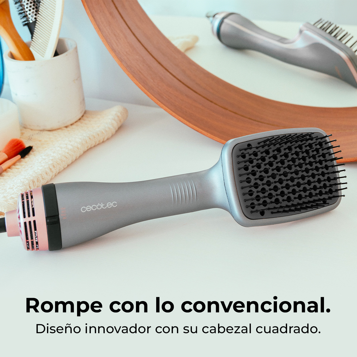 Un cepillo secador de pelo gris con cabezal cuadrado. Otros elementos incluyen un espejo, peines y brochas de maquillaje sobre una superficie blanca.