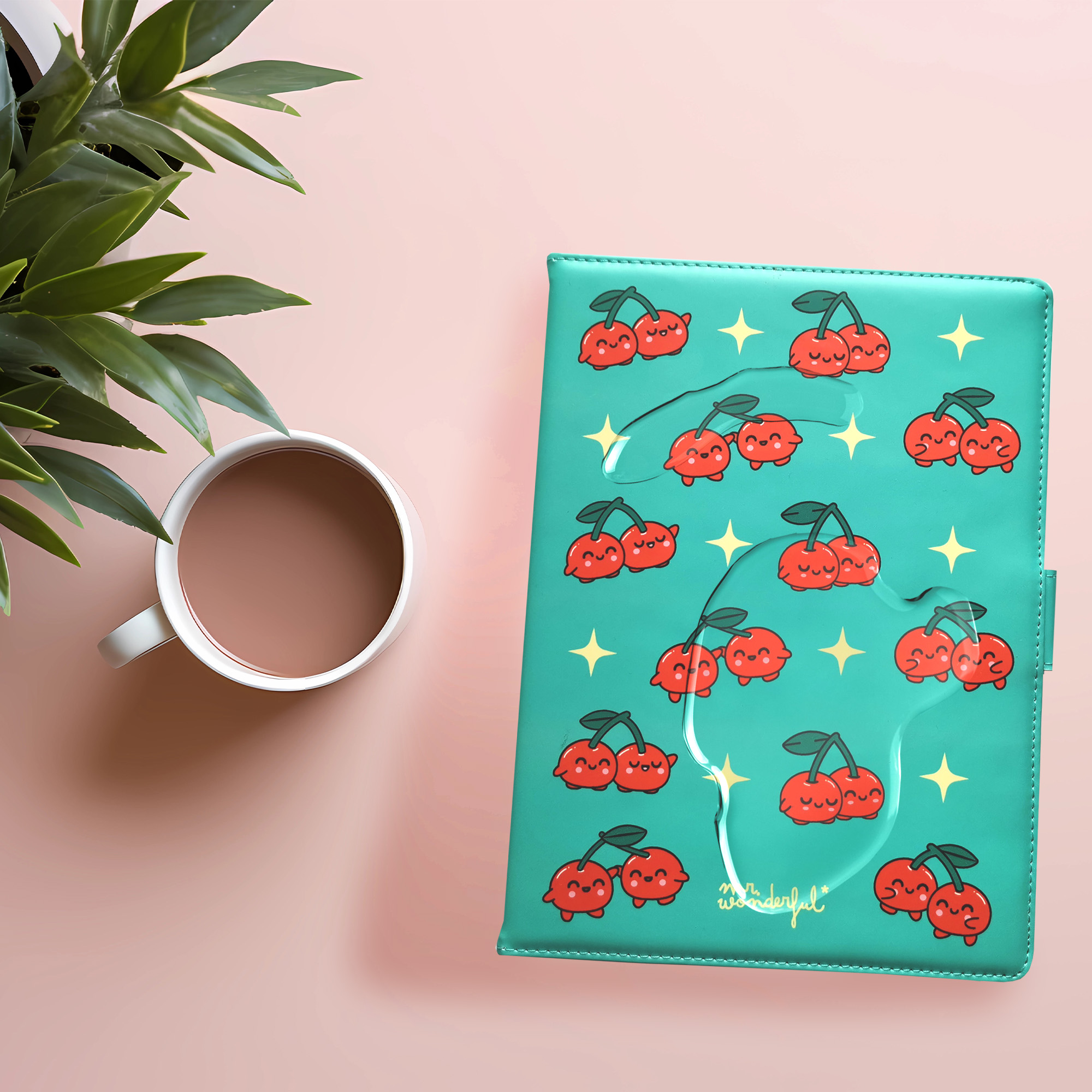 Un planificador verde azulado con diseños de cerezas, una taza de café y una planta sobre una superficie rosa.