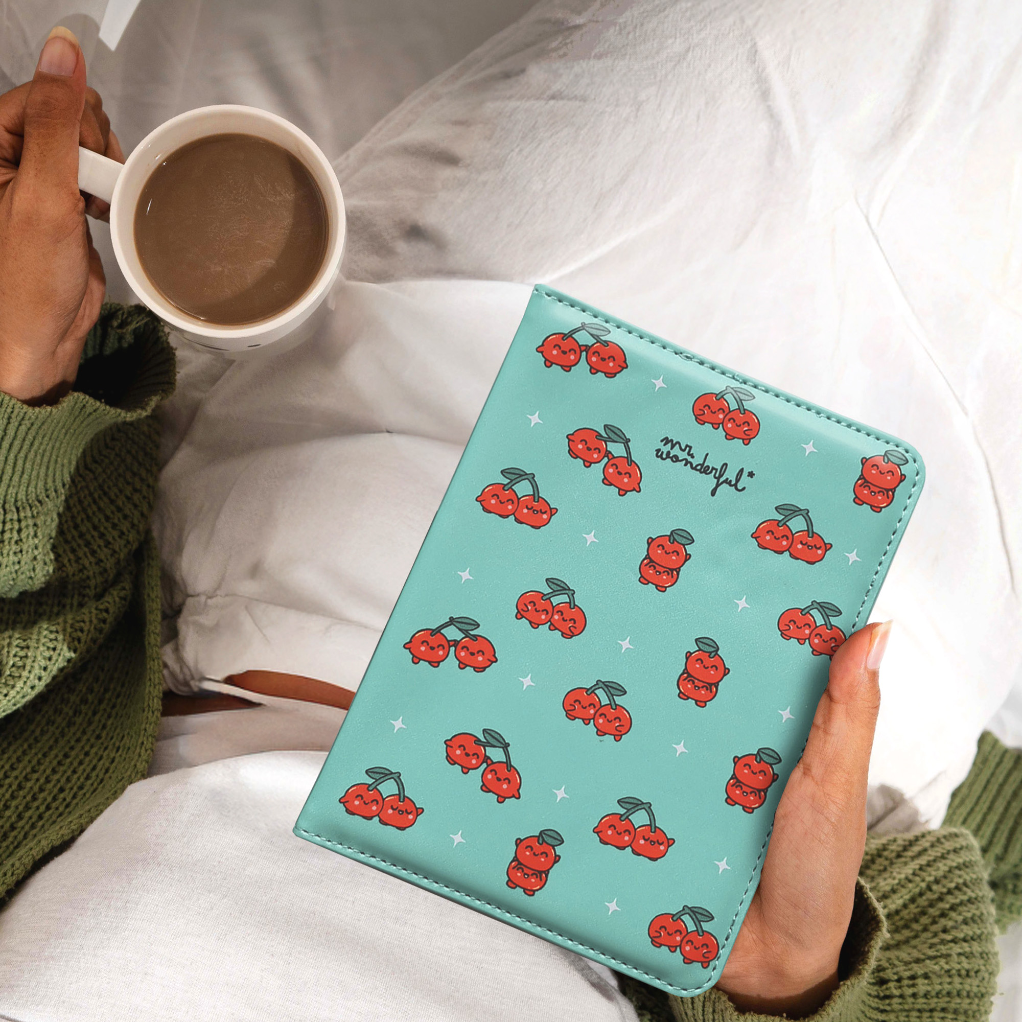 Persona sosteniendo una taza de café y un cuaderno turquesa con ilustraciones de cerezas sobre ropa de cama blanca.