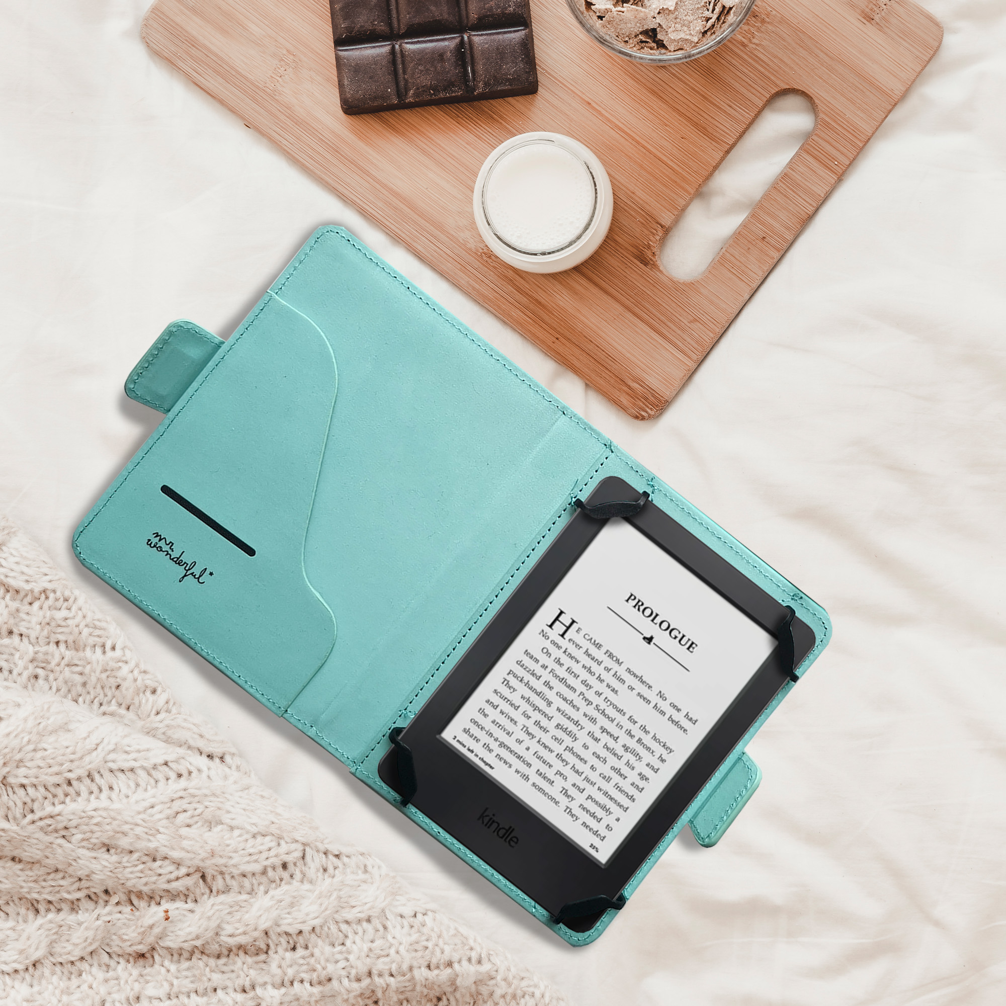 Kindle en una funda color turquesa, sobre una cama blanca, con chocolate, leche y avena. La funda dice 'mr. wonderful'.