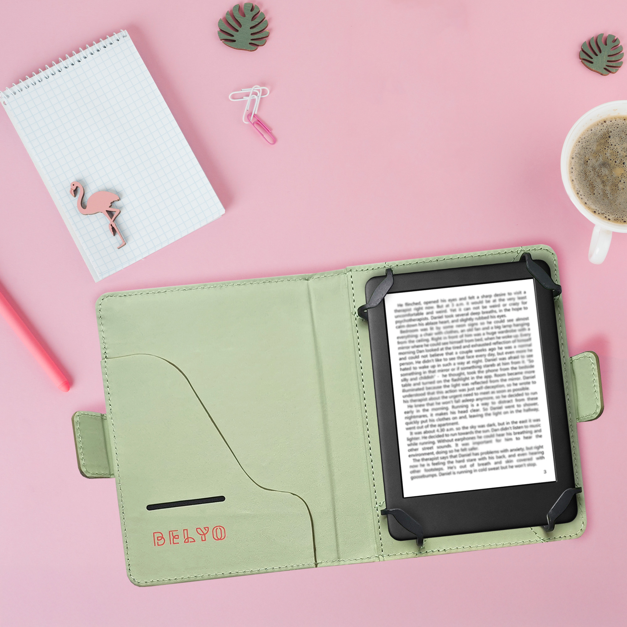 Una funda verde abierta para libros electrónicos con un libro, sobre una superficie rosa con taza de café, bloc de notas y decoración de flamenco.